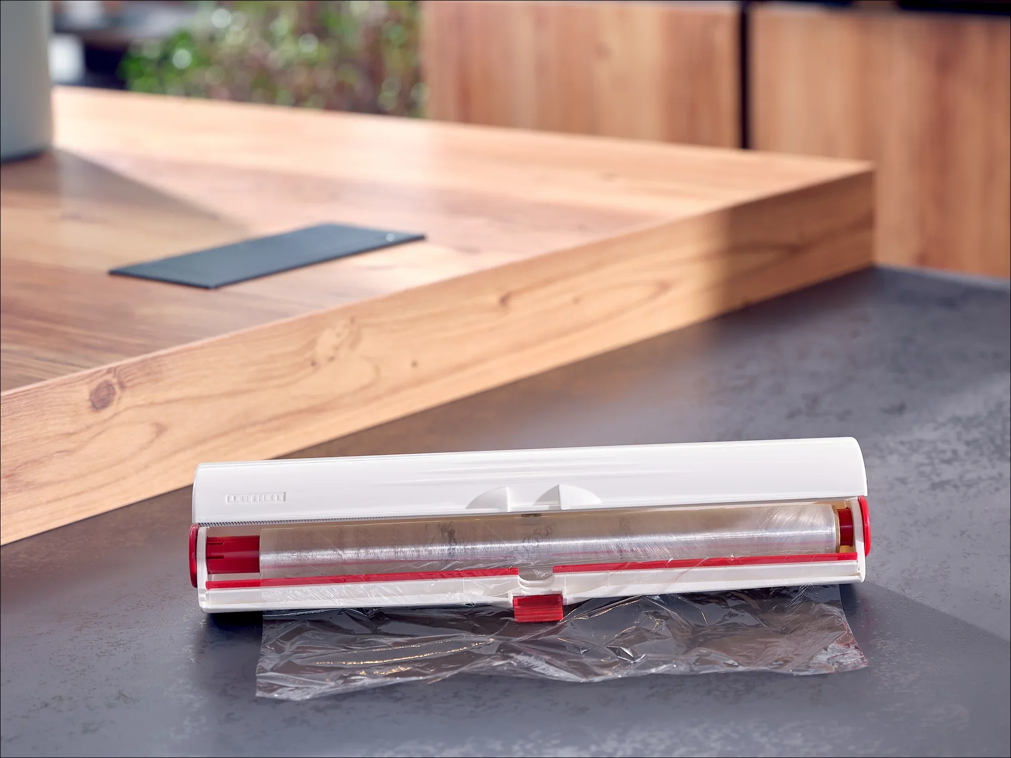 A white foil dispenser from Leifheit stands on a modern kitchen worktop. The dispenser is open and shows a pull-out plastic film.