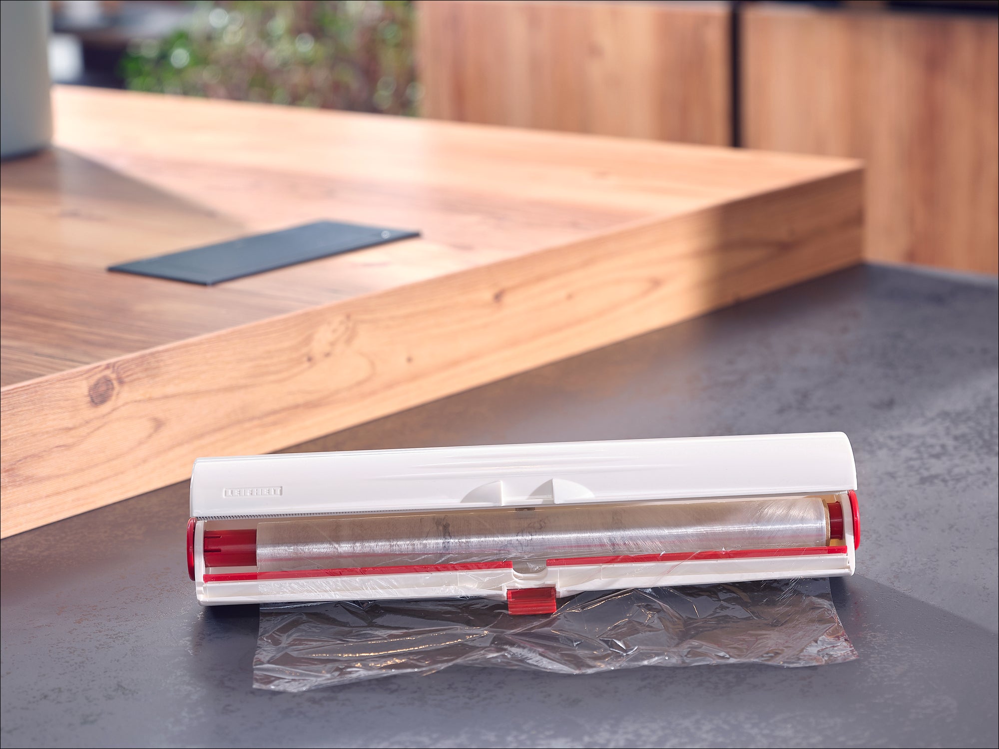 A white foil dispenser from Leifheit stands on a modern kitchen worktop. The dispenser is open and shows a pull-out plastic film.