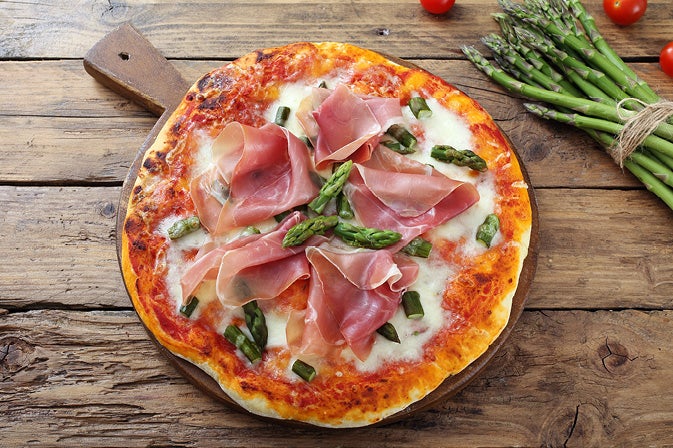 Selbstbelegte Pizza – das schnelle Frühlingsrezept für alle Selbstgemachte Pizza mit Schinken, gruenem Spargel und Kaese auf einem Holzbrett, daneben frischer Spargel und Kirschtomaten auf einem Holztisch.