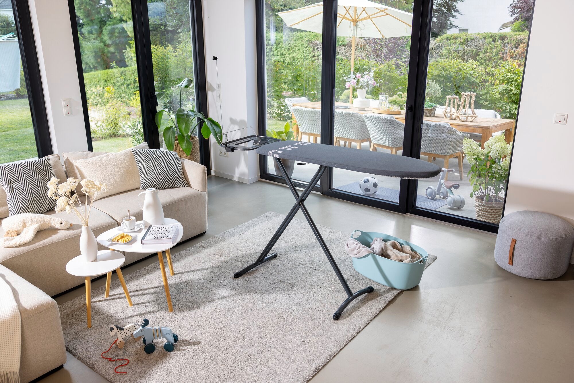 Bügelbrett Leifheit Air Board Express M Black in einem modernen Wohnzimmer mit großen Fenstern. Das Brett steht auf einem hellen Teppich, umgeben von stilvollen Möbeln und Dekorationen.