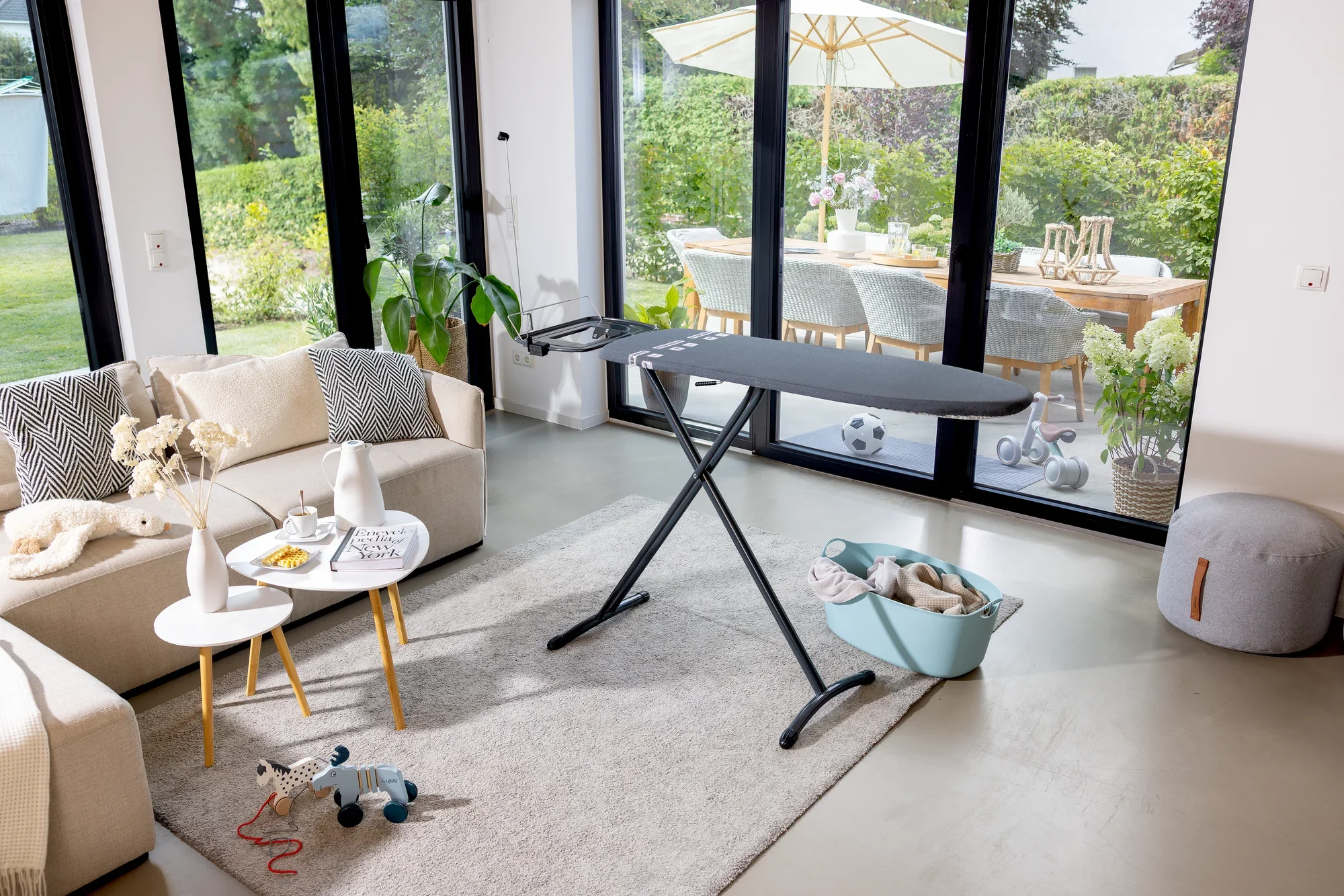 Bügelbrett Leifheit Air Board Express M Black in einem modernen Wohnzimmer mit großen Fenstern. Das Brett steht auf einem hellen Teppich, umgeben von stilvollen Möbeln und Dekorationen.
