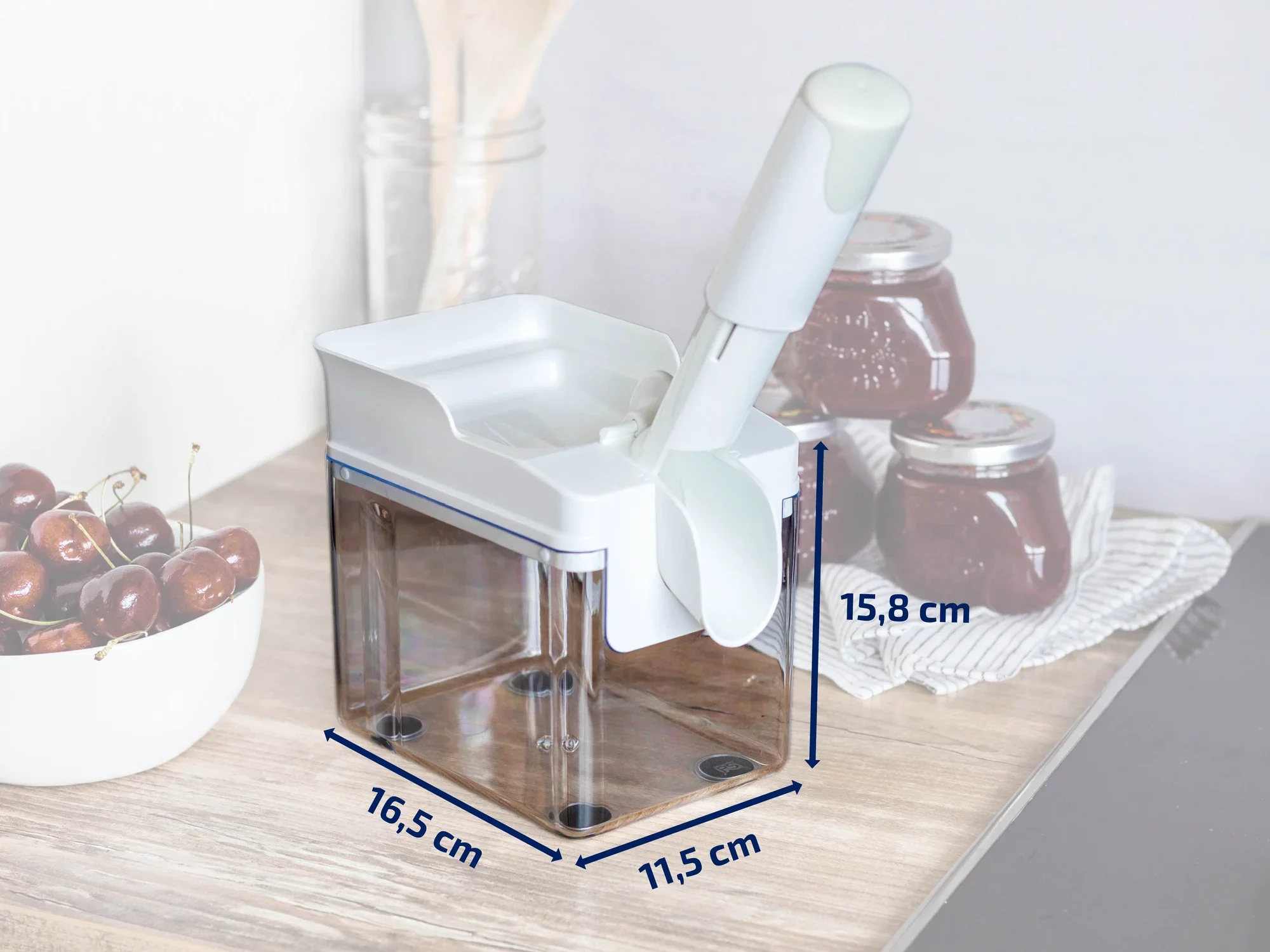 Ein weißer Kirschentkerner mit transparentem Auffangbehälter steht auf einer Küchenarbeitsplatte. Daneben befinden sich Gläser mit Marmelade. Maße: 16,5 cm x 11,5 cm x 15,8 cm.