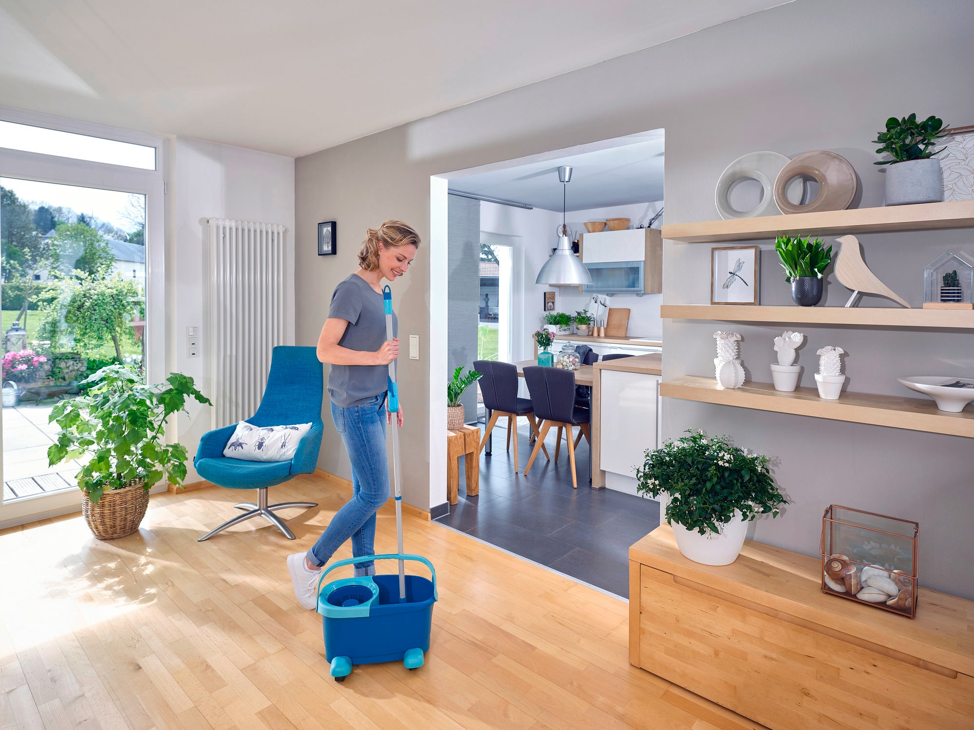 Une femme nettoie le sol en bois d'un salon moderne avec le CLEAN TWIST Disc Mop Ergo. La pièce est lumineuse et élégamment meublée, avec des plantes et des décorations.