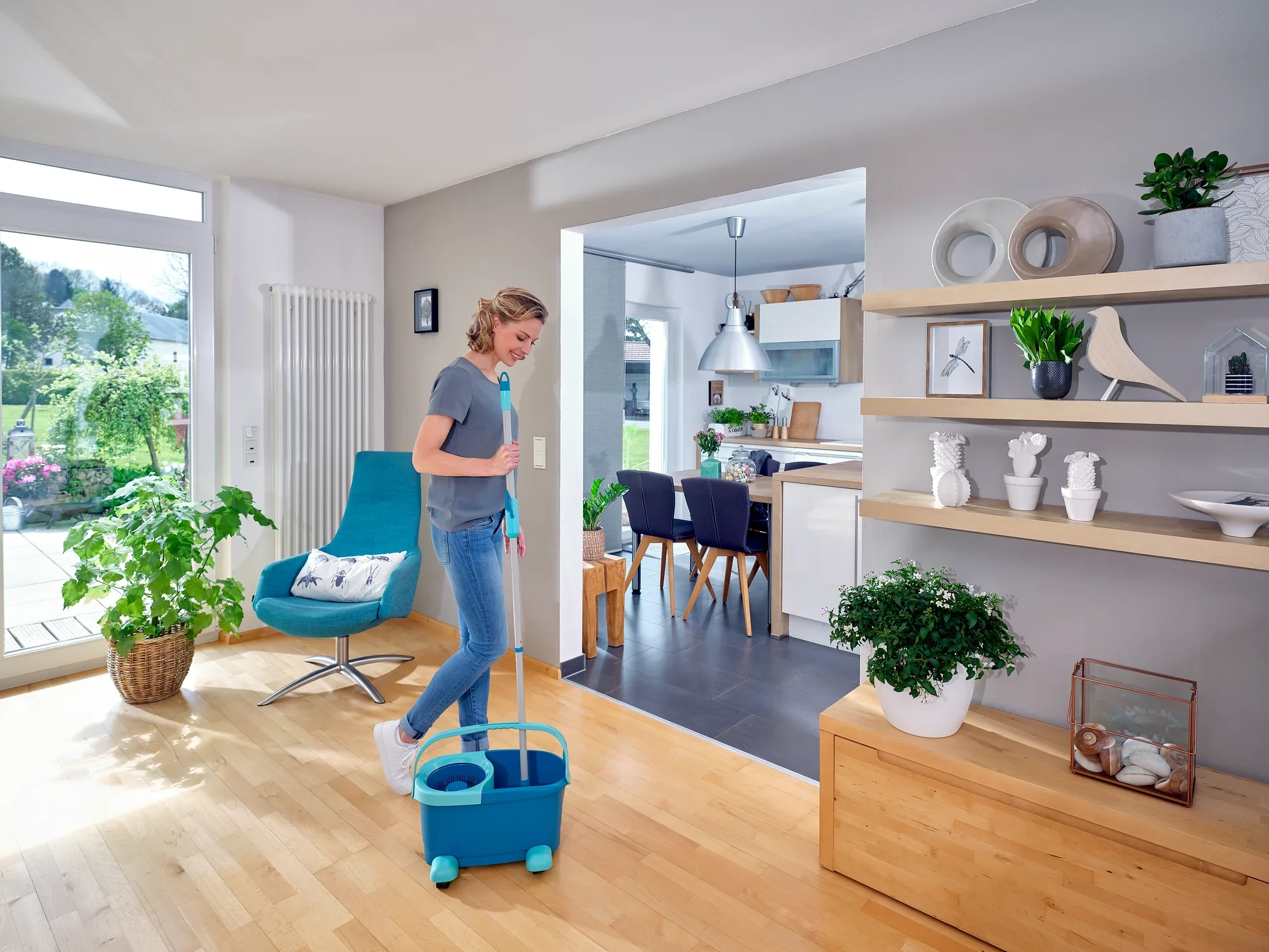Een vrouw maakt de houten vloer van een moderne woonkamer schoon met de CLEAN TWIST Disc Mop Ergo. De kamer is licht en stijlvol ingericht met planten en decoraties.