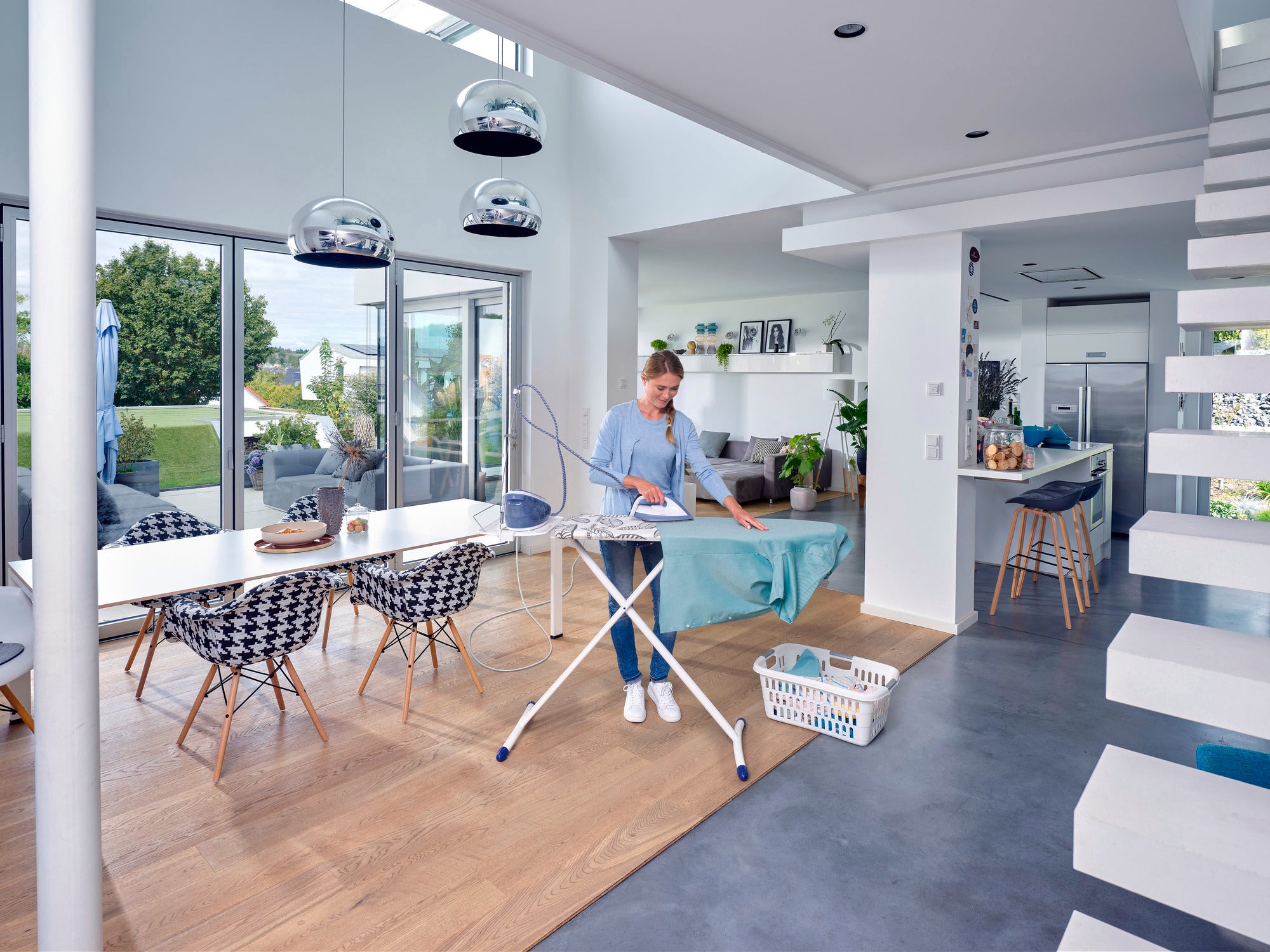 Table à repasser Air Board Express M Solid : une personne repasse dans une pièce moderne et lumineuse avec de grandes fenêtres. La table est recouverte d'un tissu bleu et un panier à linge se trouve à côté.