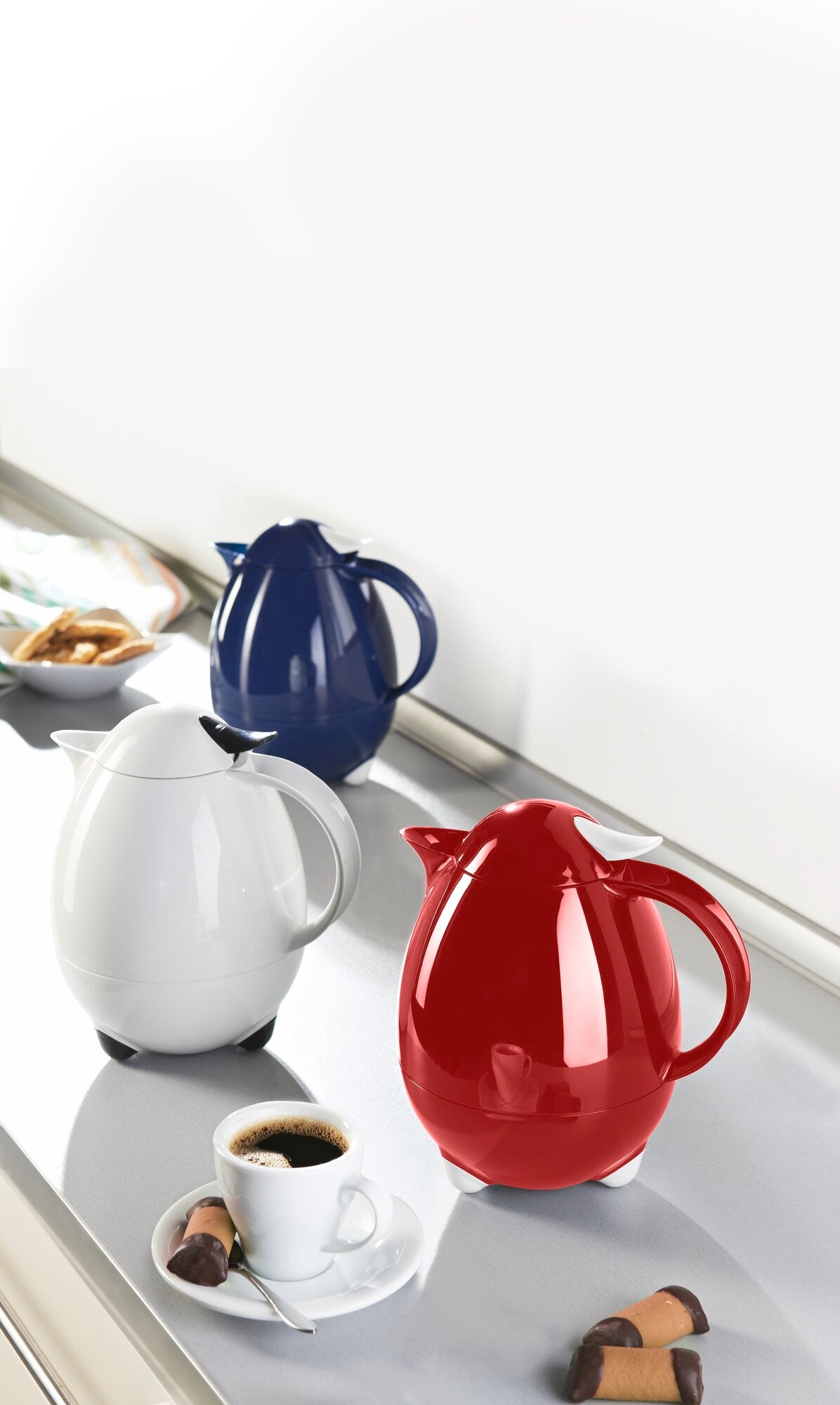Isolierkannen Columbus in Weiß, Blau und Rot auf einer Küchenarbeitsplatte. Die weiße Kanne steht neben einer Tasse Kaffee und Pralinen, ideal für stilvolle Heißgetränke.