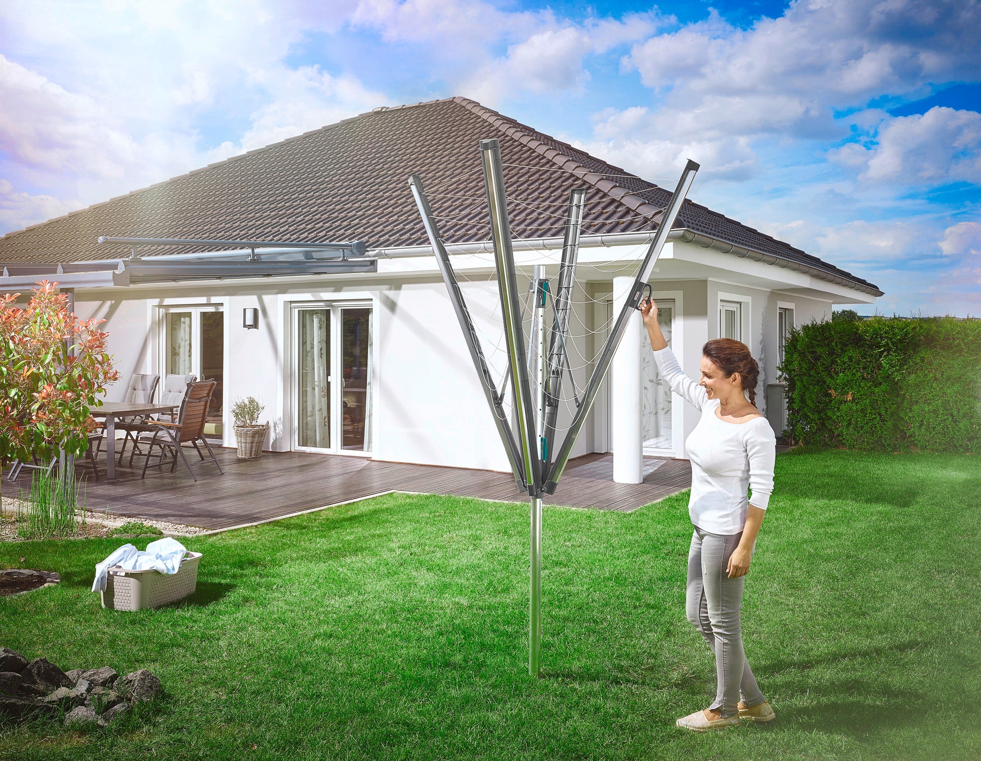 A woman stands in the garden in front of a modern detached house and uses the Linomatic 500 Plus rotary clothes dryer. The rotary clothes dryer is open and ready for hanging laundry.
