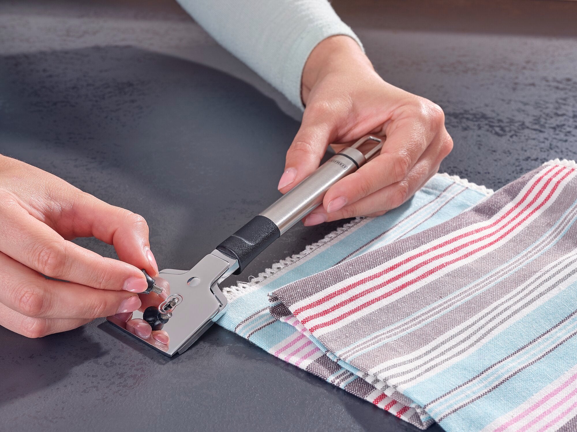 Kookplaatschraper in gebruik: Eén persoon houdt een schraper met een metalen handvat vast en maakt een oppervlak schoon. Ernaast ligt een gestreepte doek. Ideaal voor schone kookplaten.