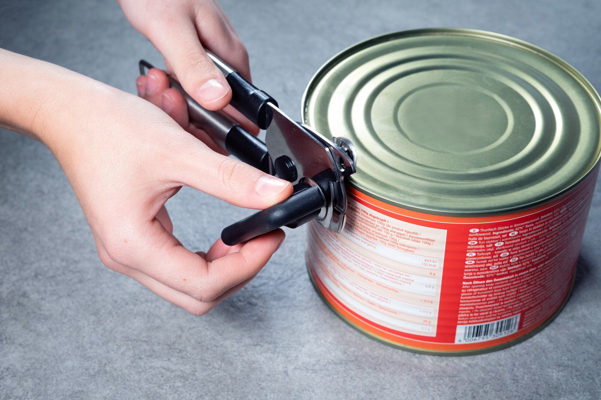 Twee handen openen een rond metalen blik met een zwarte, handmatige blikopener op een grijze ondergrond.