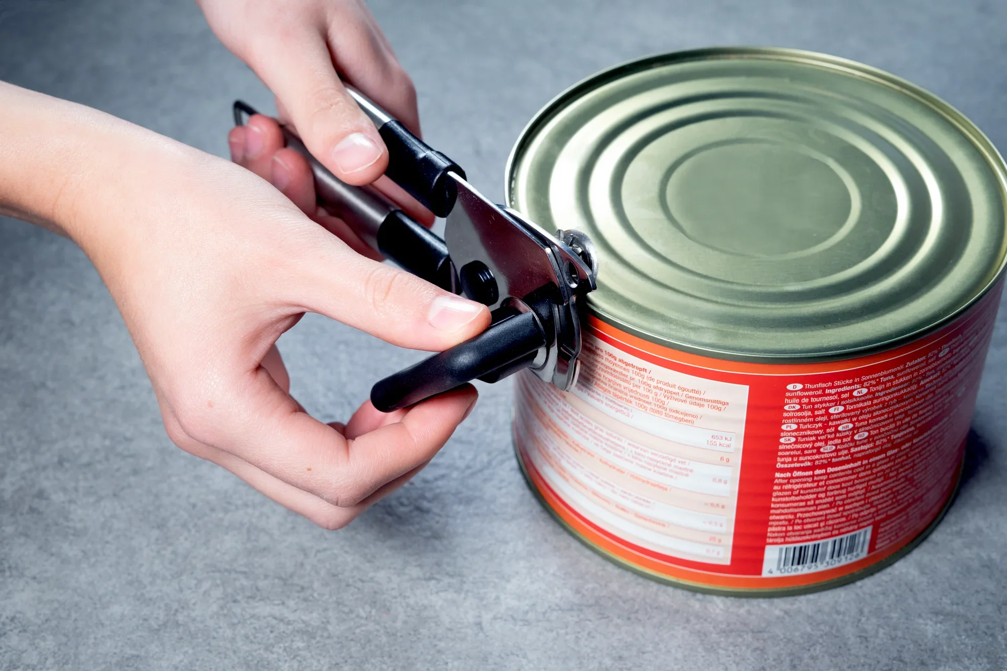 Osoba używa otwieracza do puszek, aby otworzyć puszkę. Otwieracz do puszek ma czarne uchwyty i jest umieszczany na górze puszki w celu zdjęcia wieczka.