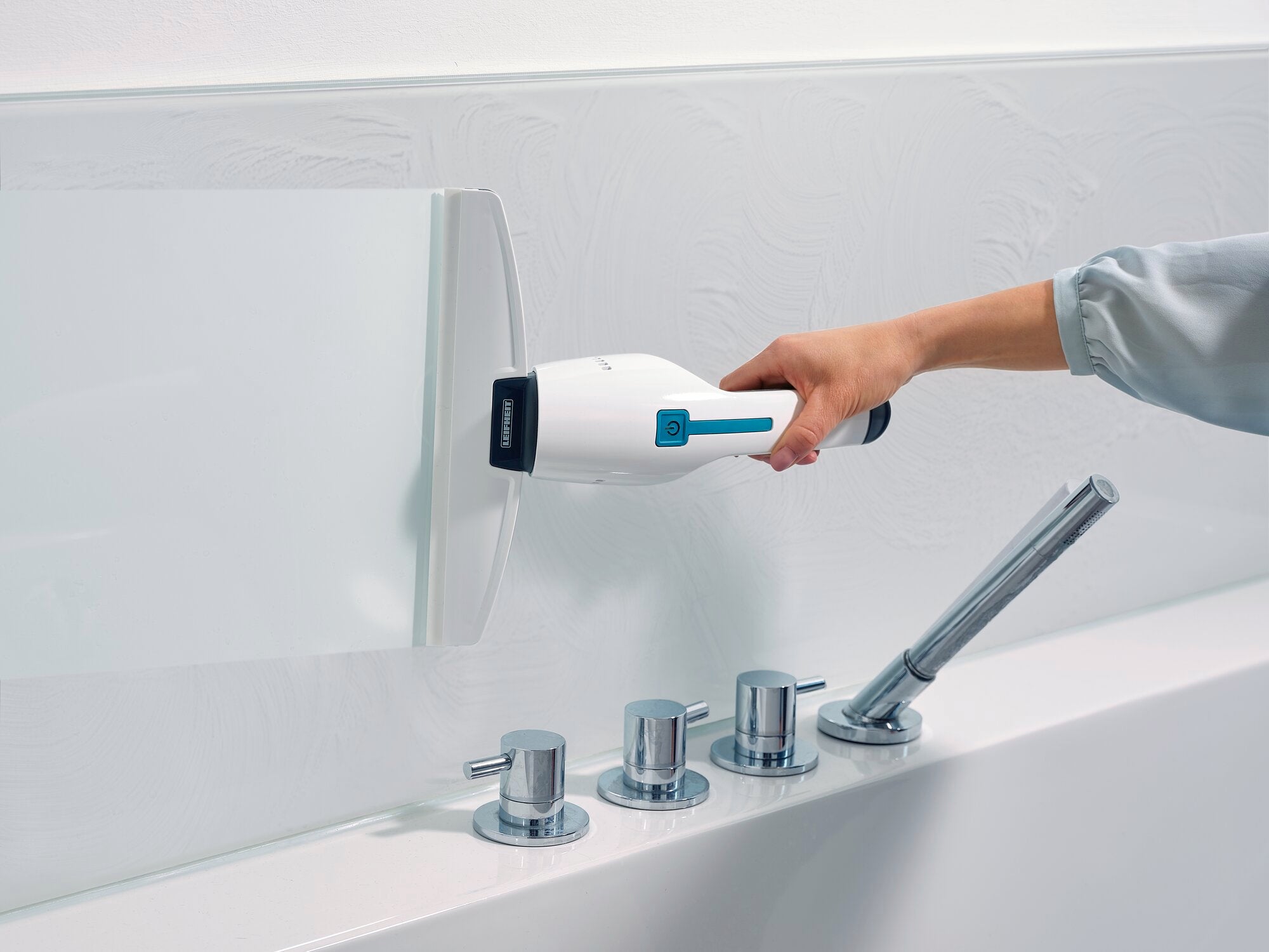 Une personne utilise l'aspirateur pour vitres et salle de bain Nemo pour nettoyer une surface en verre lisse. L'aspirateur est blanc avec des accents bleus et est utilisé dans une salle de bain moderne.