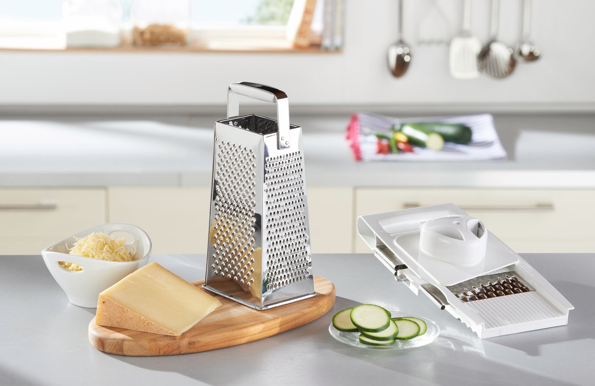 Vierkantreibe aus Edelstahl auf einer Küchenarbeitsplatte, umgeben von geriebenem Käse, einem Stück Hartkäse und geschnittenen Zucchinischeiben. Ideal zum Reiben und Schneiden.