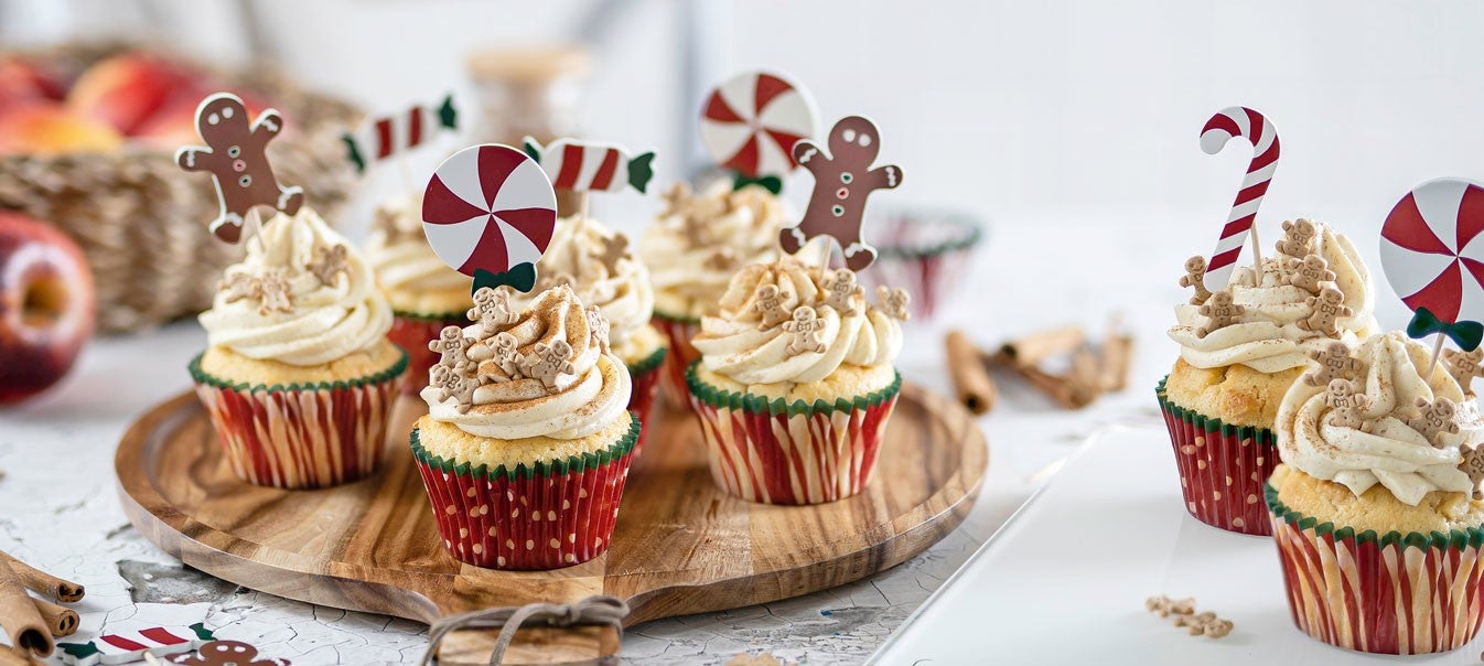 Weihnachtliche Cupcakes mit Sahnehaube, Zimt, Lebkuchenmaennchen und Zuckerstangen-Dekor auf einem Holztablett arrangiert.