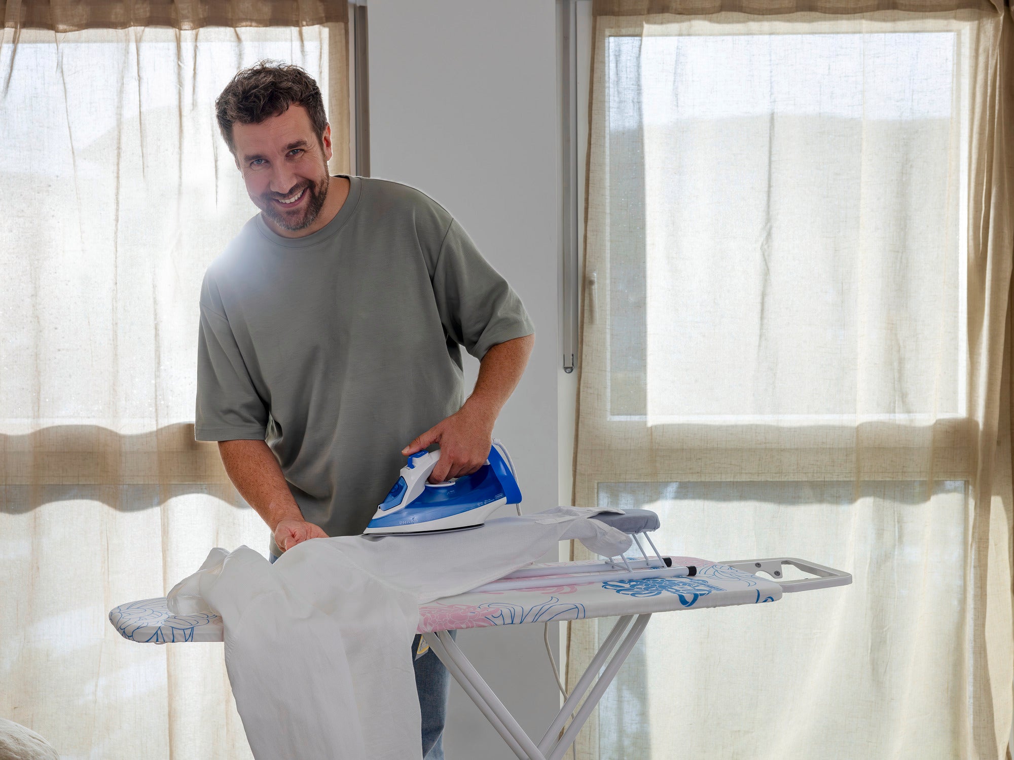 Een man strijkt een overhemd op een mouwplank voor een raam met gordijnen. Hij glimlacht en draagt een grijs T-shirt. De mouwplank heeft een kleurrijke hoes.