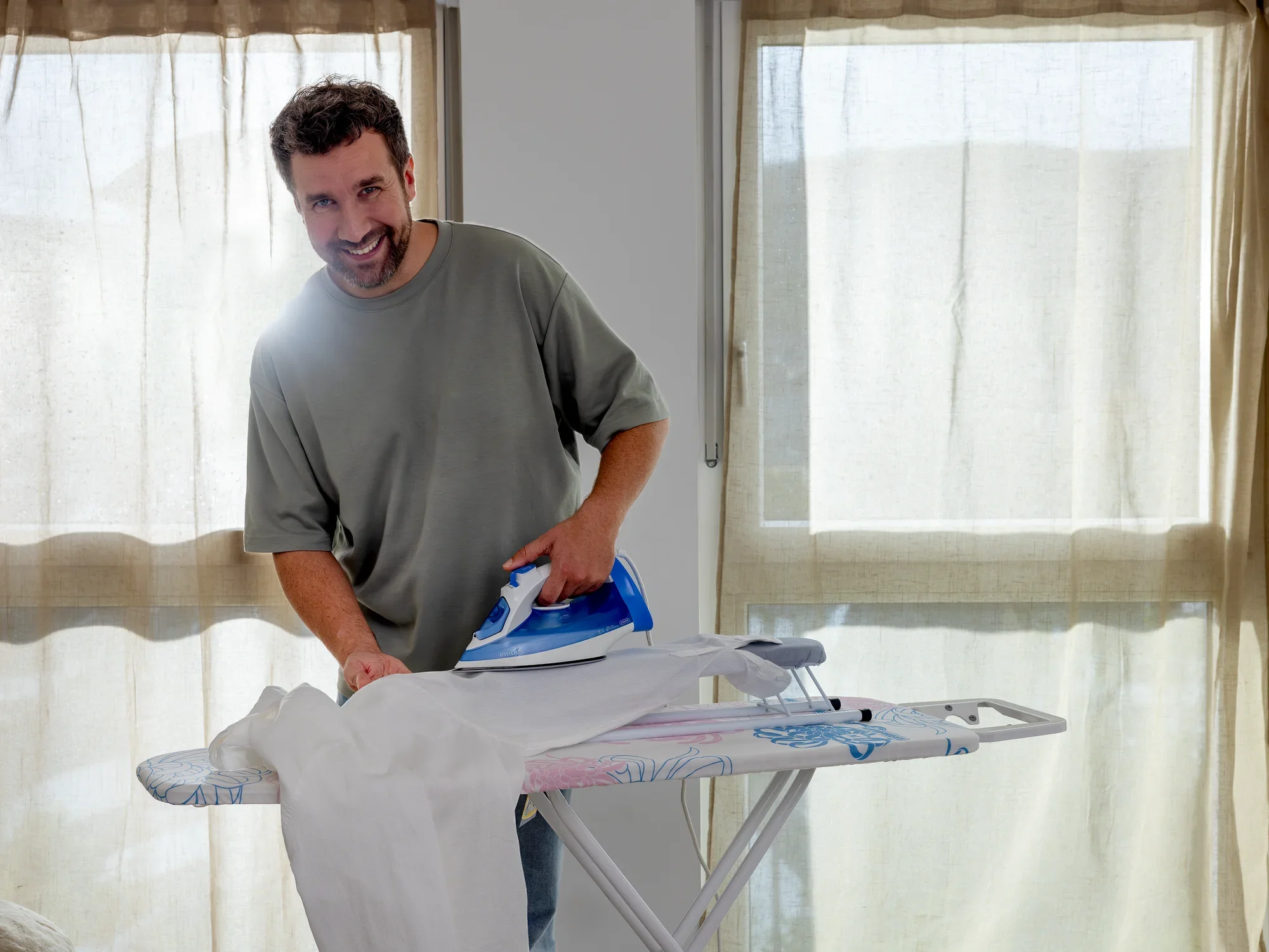 Een man strijkt een overhemd op een mouwplank voor een raam met gordijnen. Hij glimlacht en draagt een grijs T-shirt. De mouwplank heeft een kleurrijke hoes.