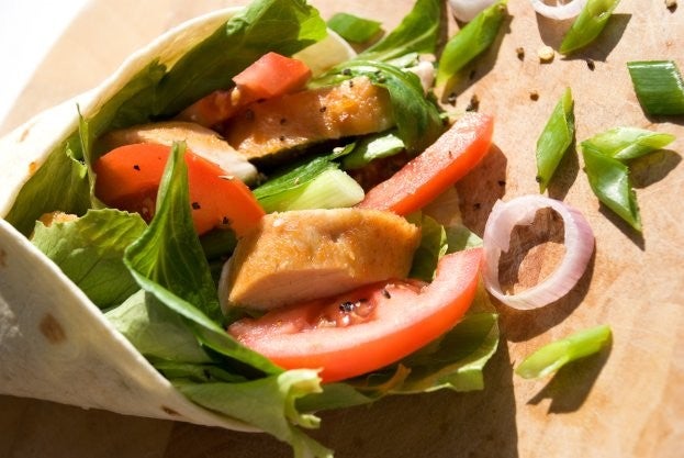 Ein Wrap gefuellt mit saftigem Haehnchen, knackigem Salat, Tomatenscheiben und Fruehlingszwiebeln auf einem hoelzernen Schneidebrett.