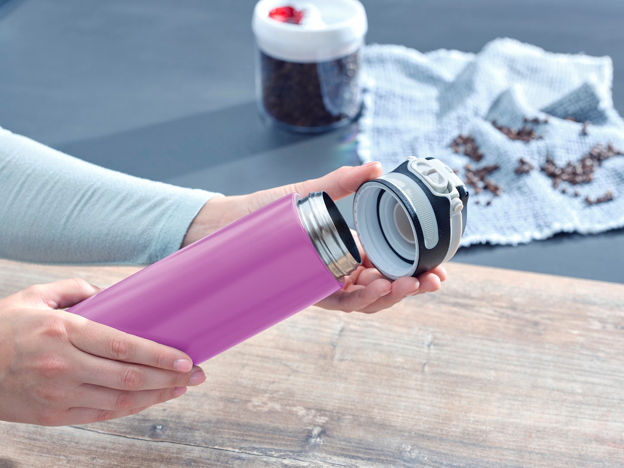 Izolowany kubek Flip 600 ml w kolorze fioletowym jest otwarty. W tle widać ziarna kawy i ściereczkę. Kubek jest idealny do podróży i utrzymuje napoje gorące lub zimne.