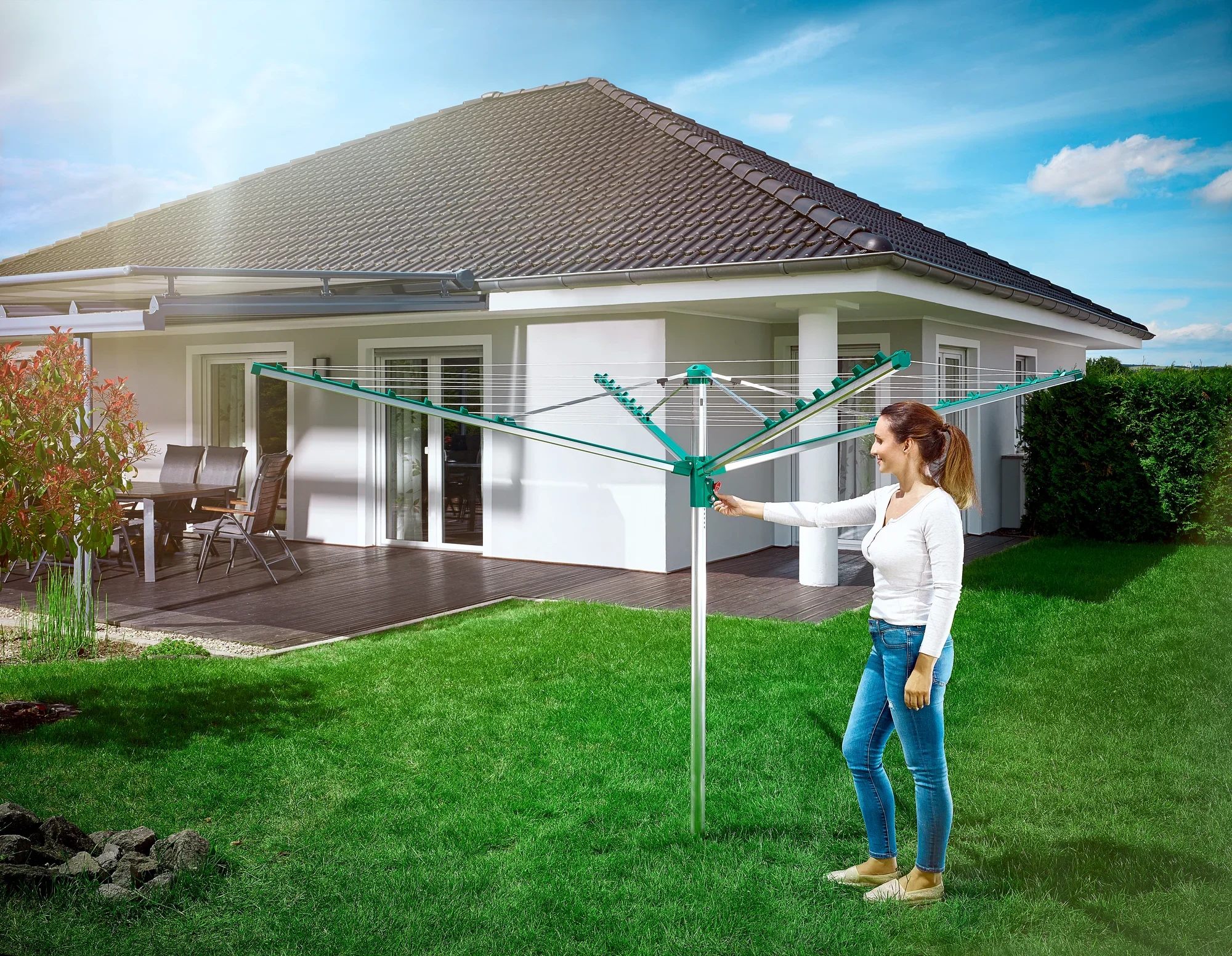 Une femme se tient dans le jardin et utilise le séchoir à linge Linomatic 400 Comfort. À l'arrière-plan, on aperçoit une maison individuelle moderne avec une terrasse. Le ciel est bleu et ensoleillé.