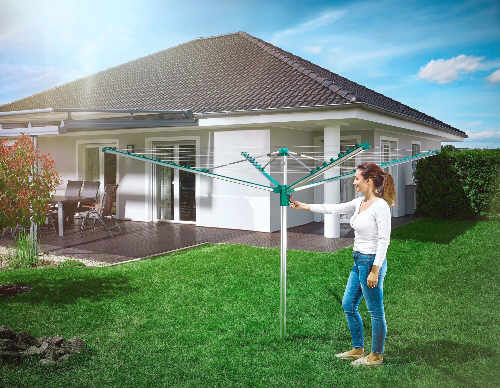 Eine Frau steht im Garten und benutzt die Wäschespinne Linomatic 400 Comfort. Im Hintergrund ist ein modernes Einfamilienhaus mit Terrasse zu sehen. Der Himmel ist blau und sonnig.
