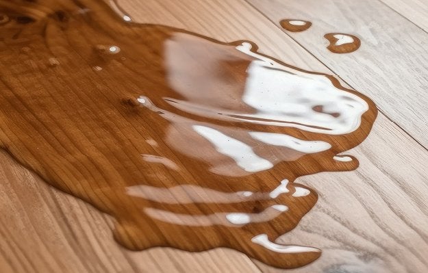 Holzboden mit verschuettetem Wasser, das sich unregelmaessig ausbreitet und reflektiert; es bildet glaenzende, fluessige Pfuetzen.
