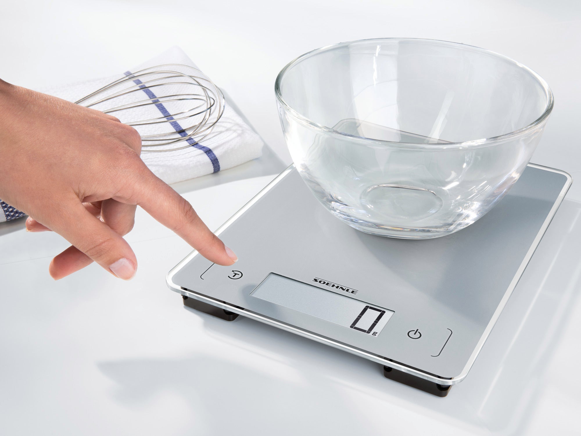 Une balance de cuisine numérique avec surface en verre est utilisée. Une main pointe vers l'écran tandis qu'un bol en verre est posé sur la balance. Un fouet est posé à l'arrière-plan.