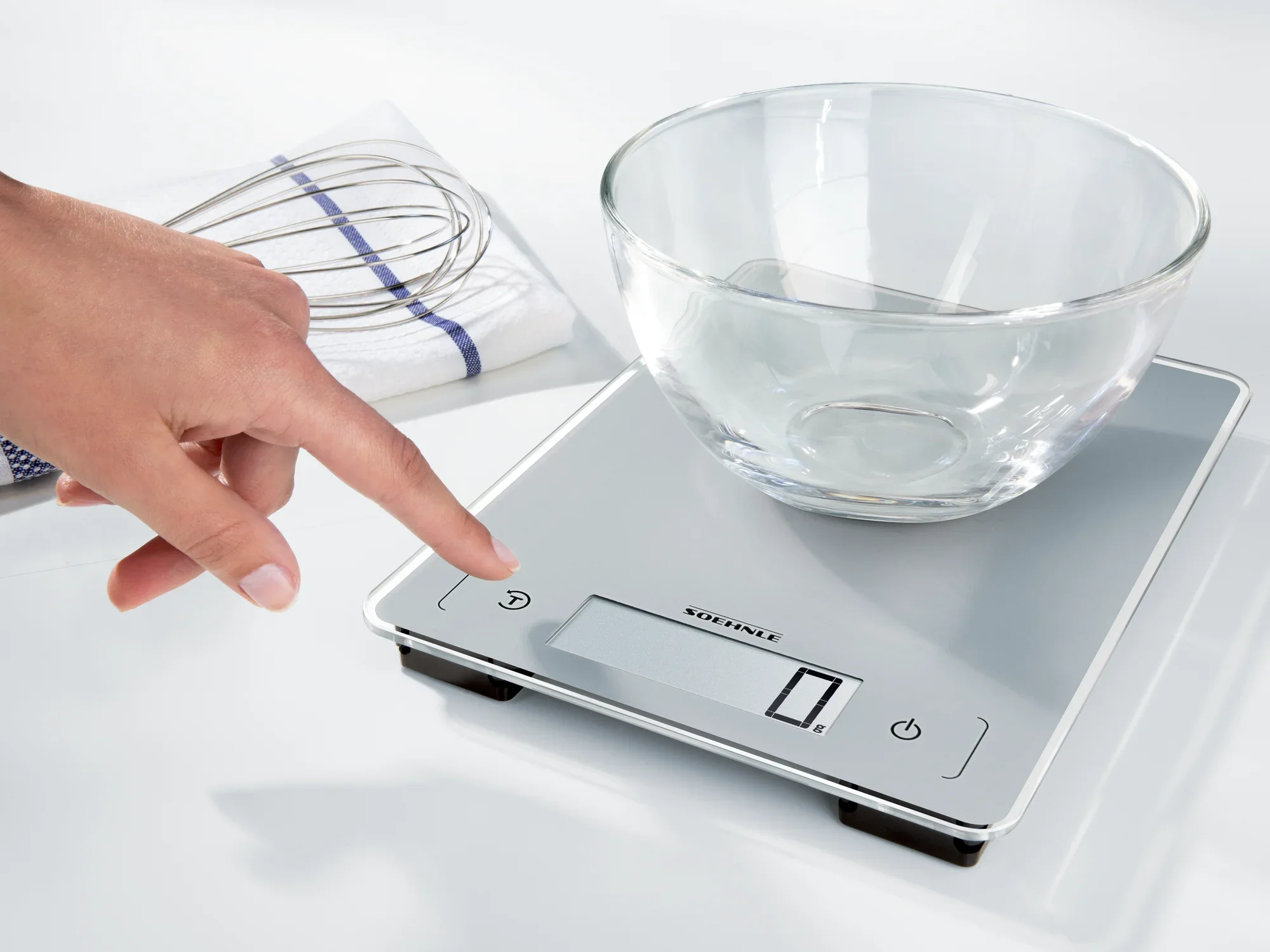 Eine digitale Küchenwaage mit Glasoberfläche wird verwendet. Eine Hand zeigt auf das Display, während eine Glasschüssel auf der Waage steht. Im Hintergrund liegt ein Schneebesen.
