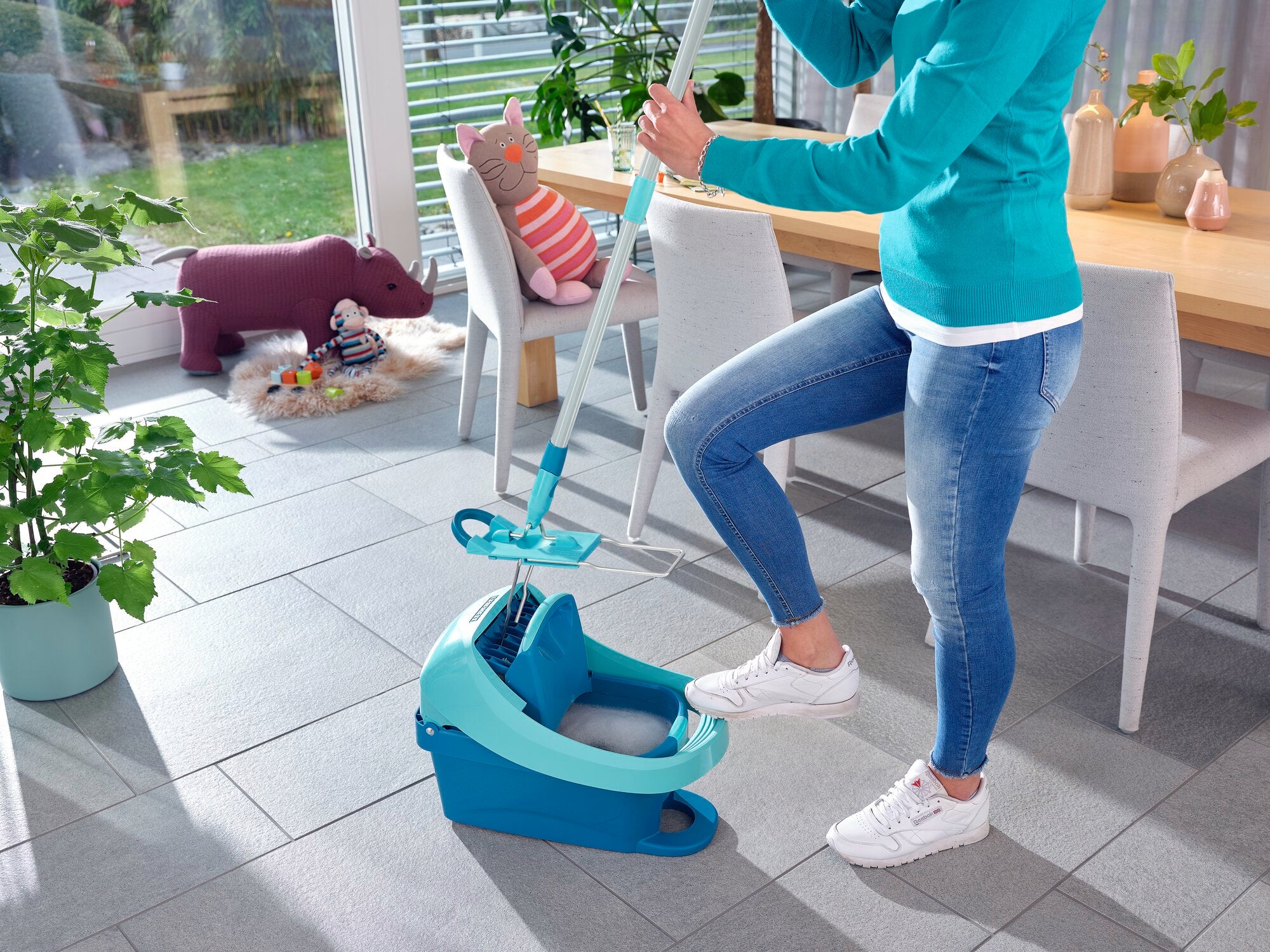 Eine Person benutzt die Wischtuchpresse Profi XL in einem modernen Wohnzimmer. Die Presse ist türkis und wird mit einem Fußpedal bedient. Im Hintergrund sind Pflanzen und ein Tisch zu sehen.