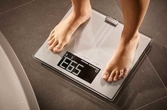 Person steht barfuss auf einer digitalen Waage mit grauer Glasflaeche, die ein Gewicht von 59,3 kg anzeigt, auf dunklem Boden.