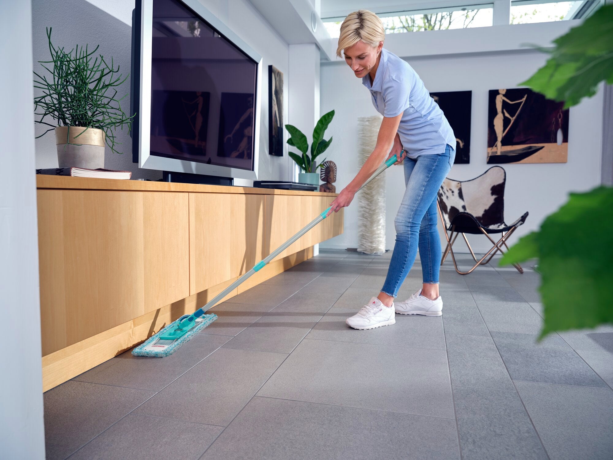 Una mujer limpia el suelo de un salón moderno con la mopa Profi XL super soft. La habitación es luminosa y está decorada con estilo, con plantas y obras de arte en las paredes.
