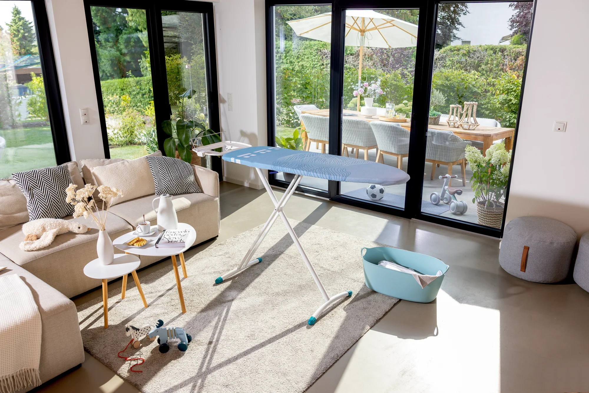 Bügelbrett Leifheit Air Board Express M Solid in einem modernen Wohnzimmer mit großen Fenstern. Das Brett steht auf einem Teppich, umgeben von stilvollen Möbeln und Dekorationen.