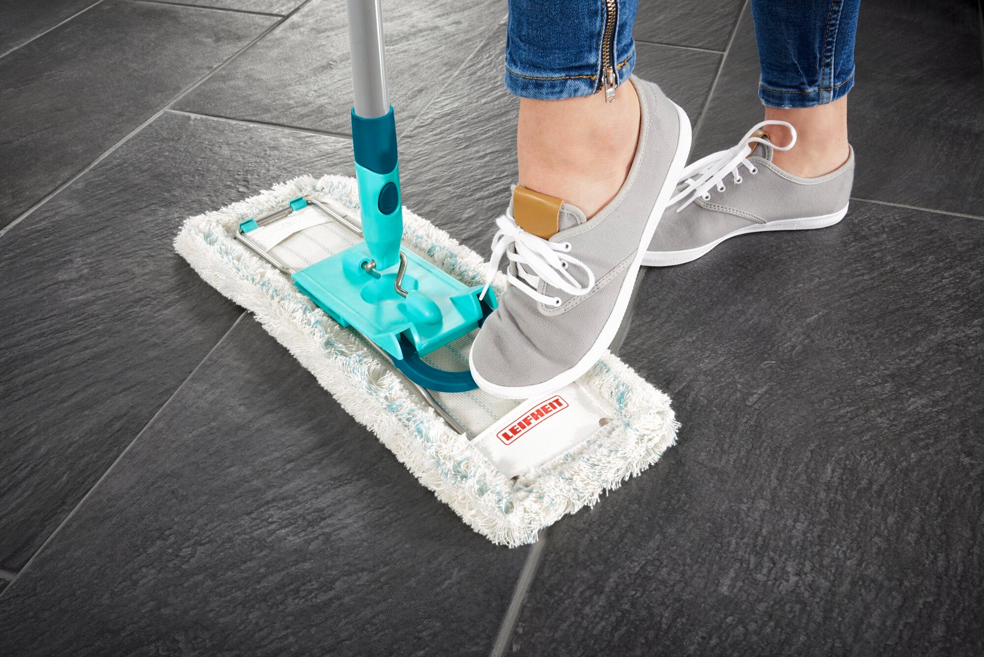 Ein Bodenwischer Profi XL wird auf dunklen Fliesen verwendet. Eine Person mit grauen Sneakers betätigt den Wischmechanismus mit dem Fuß. Ideal für gründliche Reinigung.