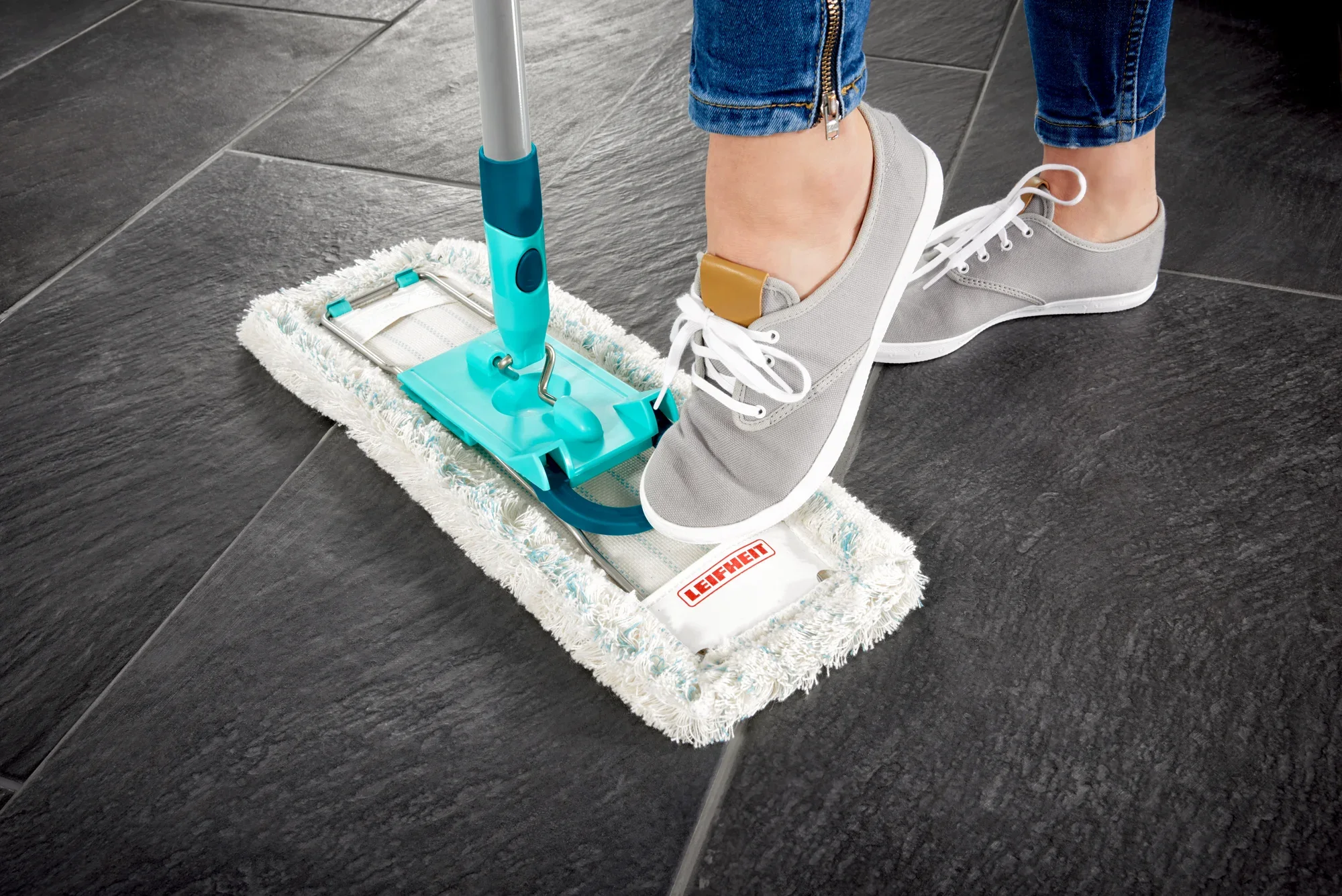 Ein Bodenwischer Profi XL wird auf dunklen Fliesen verwendet. Eine Person mit grauen Sneakers betätigt den Wischmechanismus mit dem Fuß. Ideal für gründliche Reinigung.