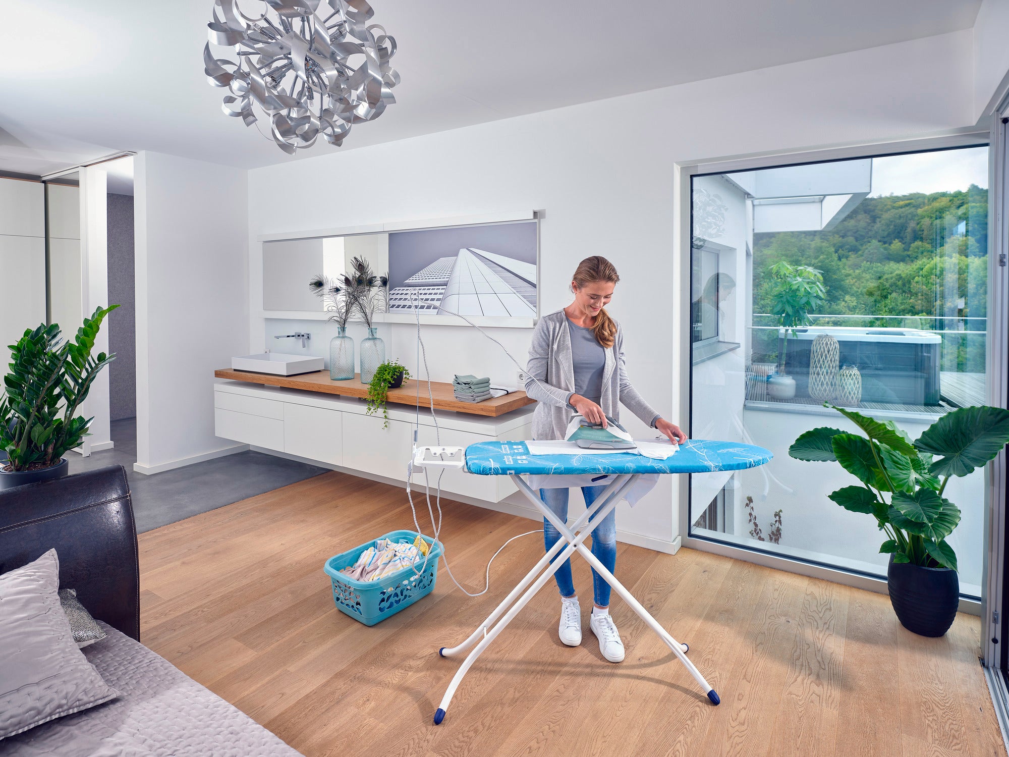 Eine Frau bügelt Kleidung auf dem Bügeltisch Air Board M Compact Plus in einem modernen, hellen Raum mit großen Fenstern und Pflanzen. Der Tisch ist leicht und funktional.