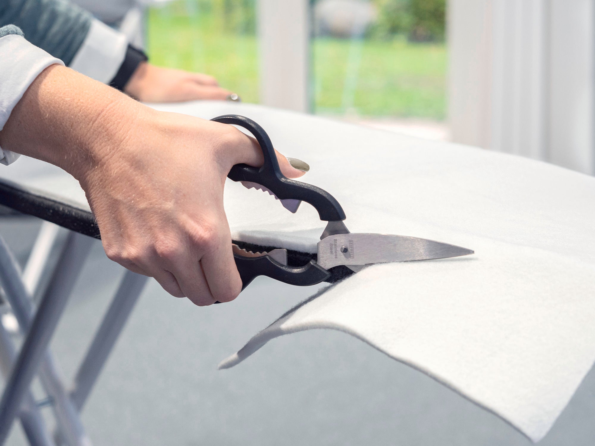 Eine Person schneidet mit einer Schere ein Bügeltischpolster auf einem Bügelbrett zu. Das Bild zeigt den präzisen Einsatz der Schere, um das Polster passend zuzuschneiden.