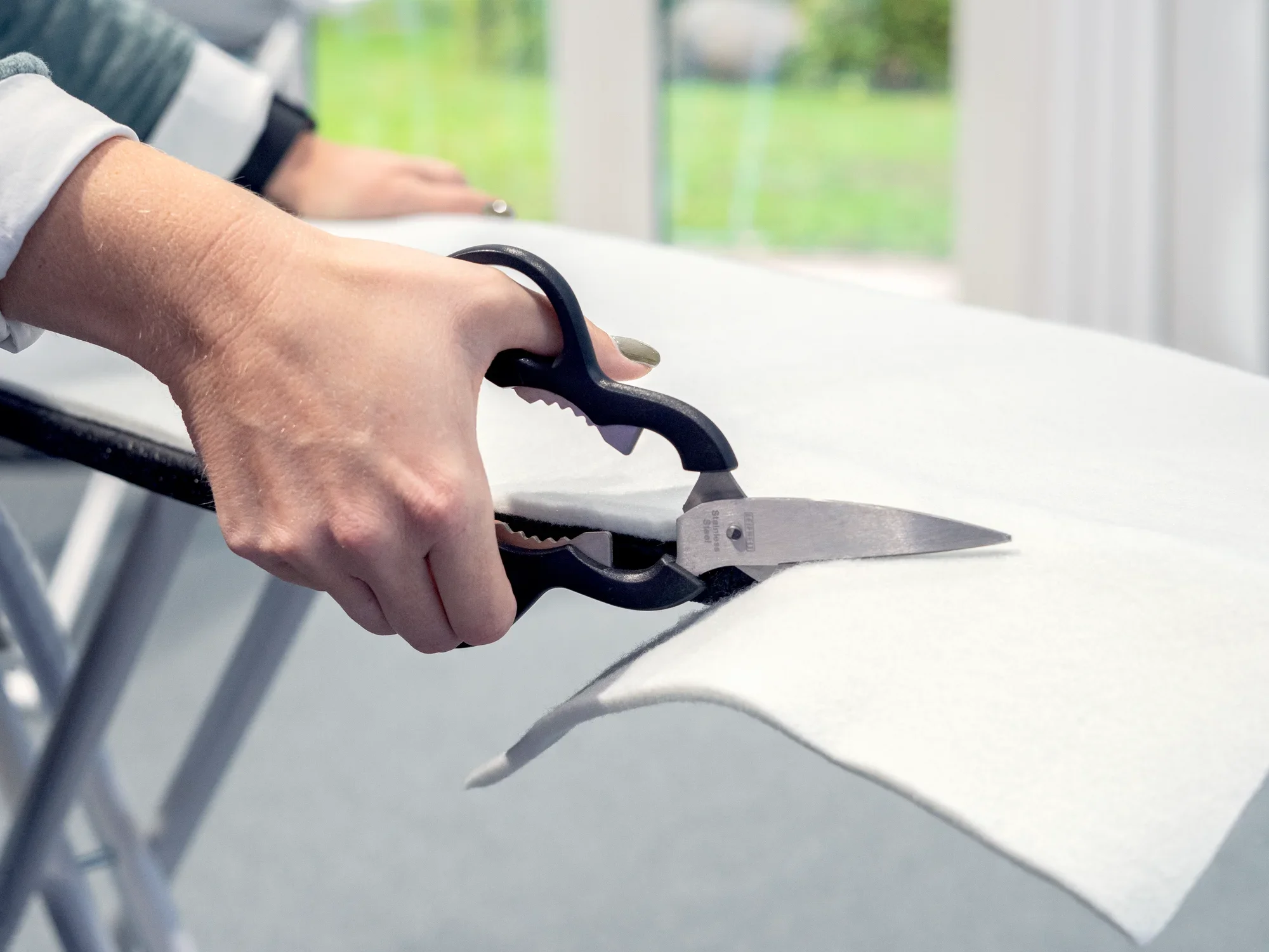Una persona recorta con unas tijeras un acolchado para tabla de planchar sobre una tabla de planchar. La imagen muestra el uso preciso de las tijeras para recortar el acolchado a la medida adecuada.