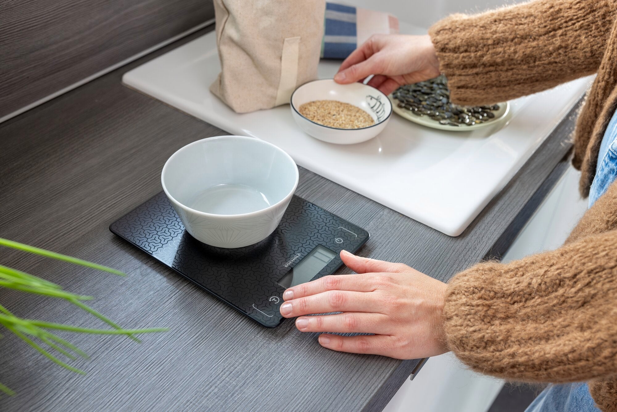Comfort Mosaic digital kitchen scales in use. A person weighs ingredients in a bowl on the scales. The scales have a modern design and an easy-to-read display.