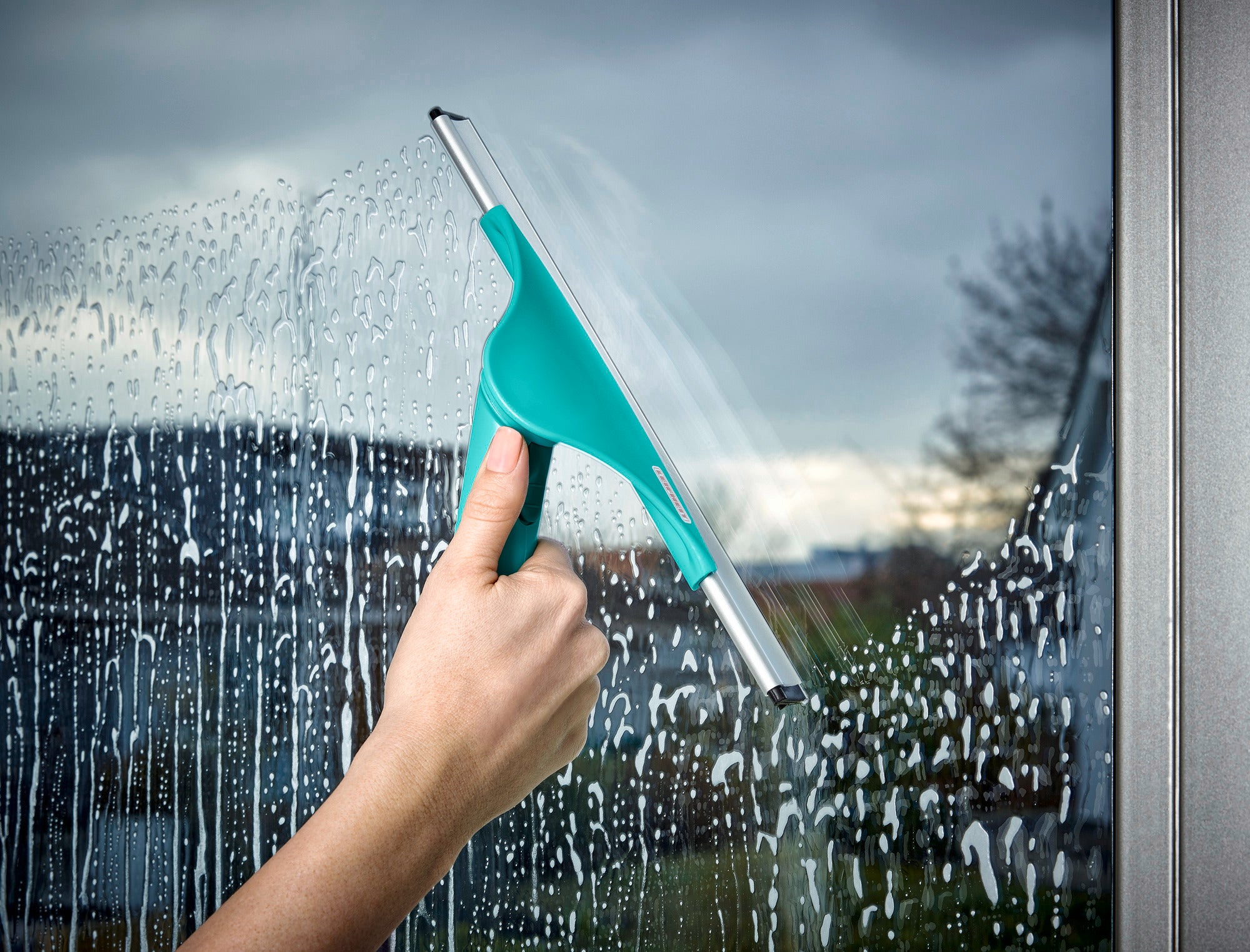 Eine Hand hält den Fensterabzieher Window Slider M und reinigt ein nasses Fenster. Der Abzieher hat einen türkisfarbenen Griff und eine Metallklinge, die das Wasser effizient entfernt.