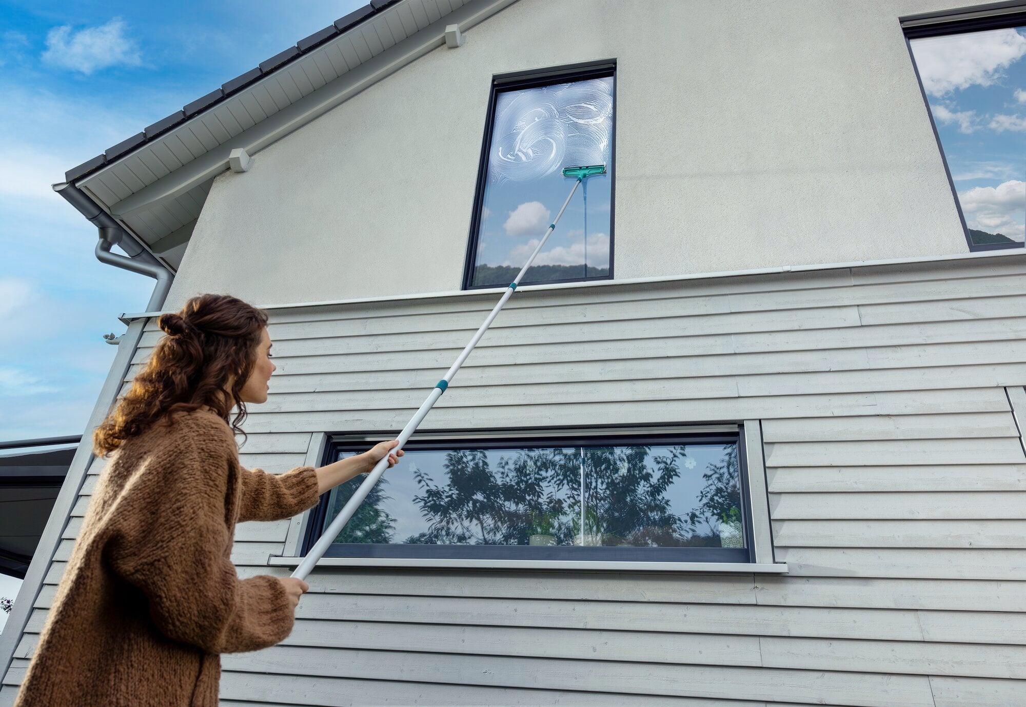 Eine Frau reinigt mit einem Teleskopstiel, der von 145 bis 400 cm verstellbar ist, ein hohes Fenster eines modernen Hauses. Der Stiel ermöglicht das Erreichen schwer zugänglicher Stellen.