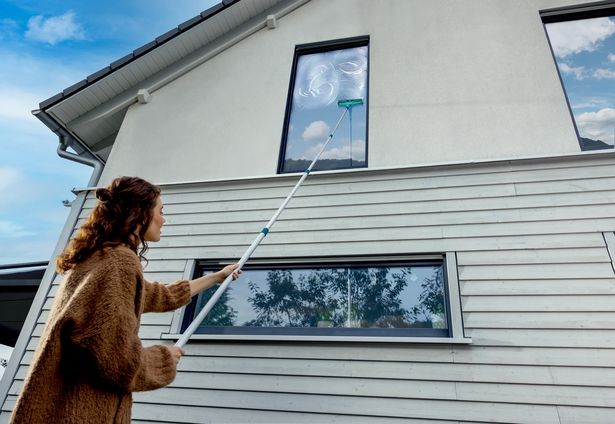 Kobieta myje wysokie okno w nowoczesnym domu za pomocą trzonka teleskopowego, który można regulować w zakresie od 145 do 400 cm. Uchwyt umożliwia dotarcie do trudno dostępnych miejsc.