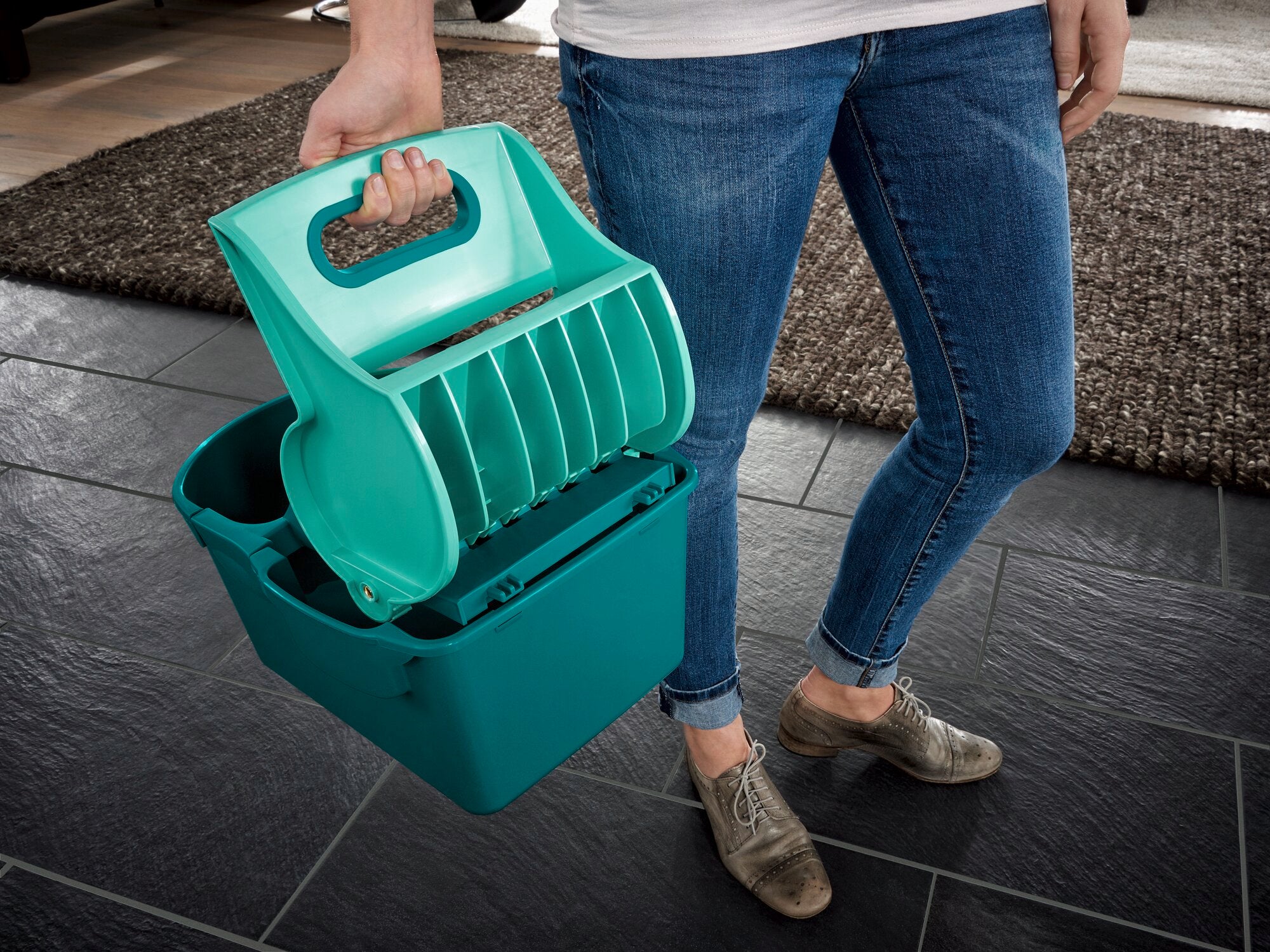 Eine Person hält die Wischtuchpresse Profi Compact in der Hand. Das Gerät ist türkis und wird auf einem dunklen Fliesenboden verwendet. Ideal für effiziente Reinigung.