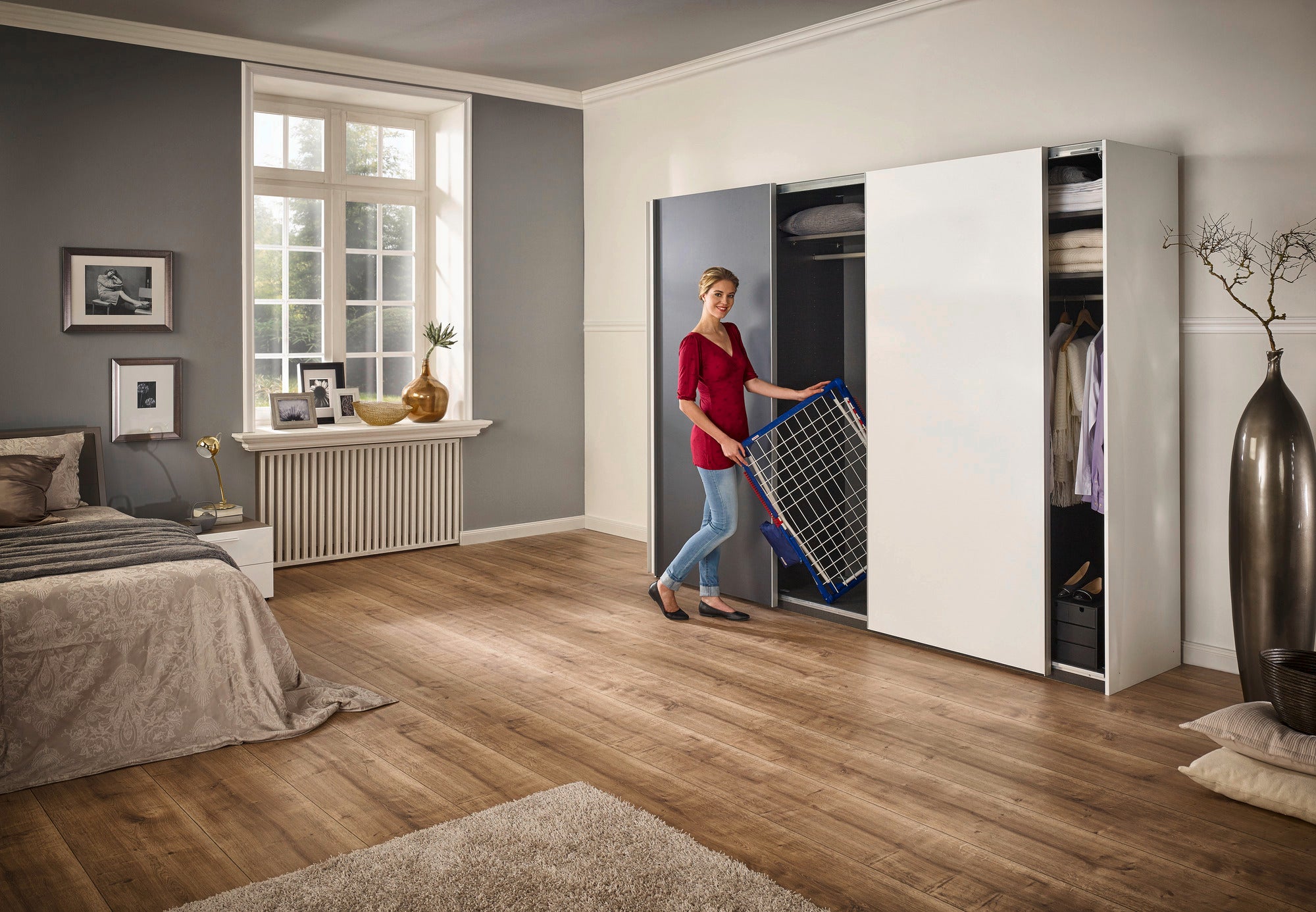 Eine Frau steht in einem modernen Schlafzimmer und verstaut den Standtrockner Pegasus 180 Solid Plus in einem weißen Schrank. Der Raum ist stilvoll eingerichtet mit Holzboden und großen Fenstern.