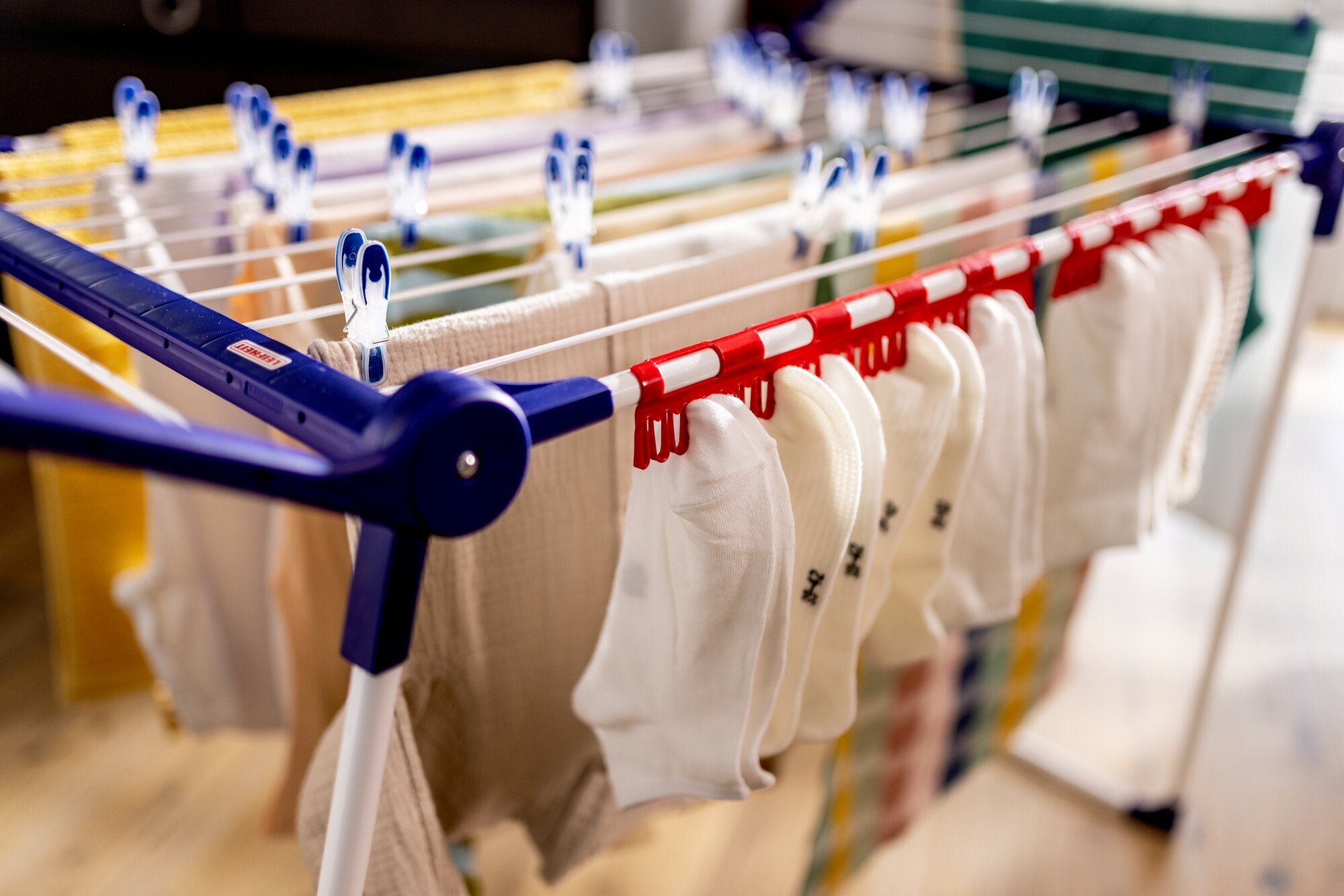 Leifheit Pegasus Family 18m Wäscheständer mit ausklappbaren Flügeln und Sockentrocknung: mehrere weiße Socken hängen an roten und blauen Klammern auf den Streben im Innenbereich.
