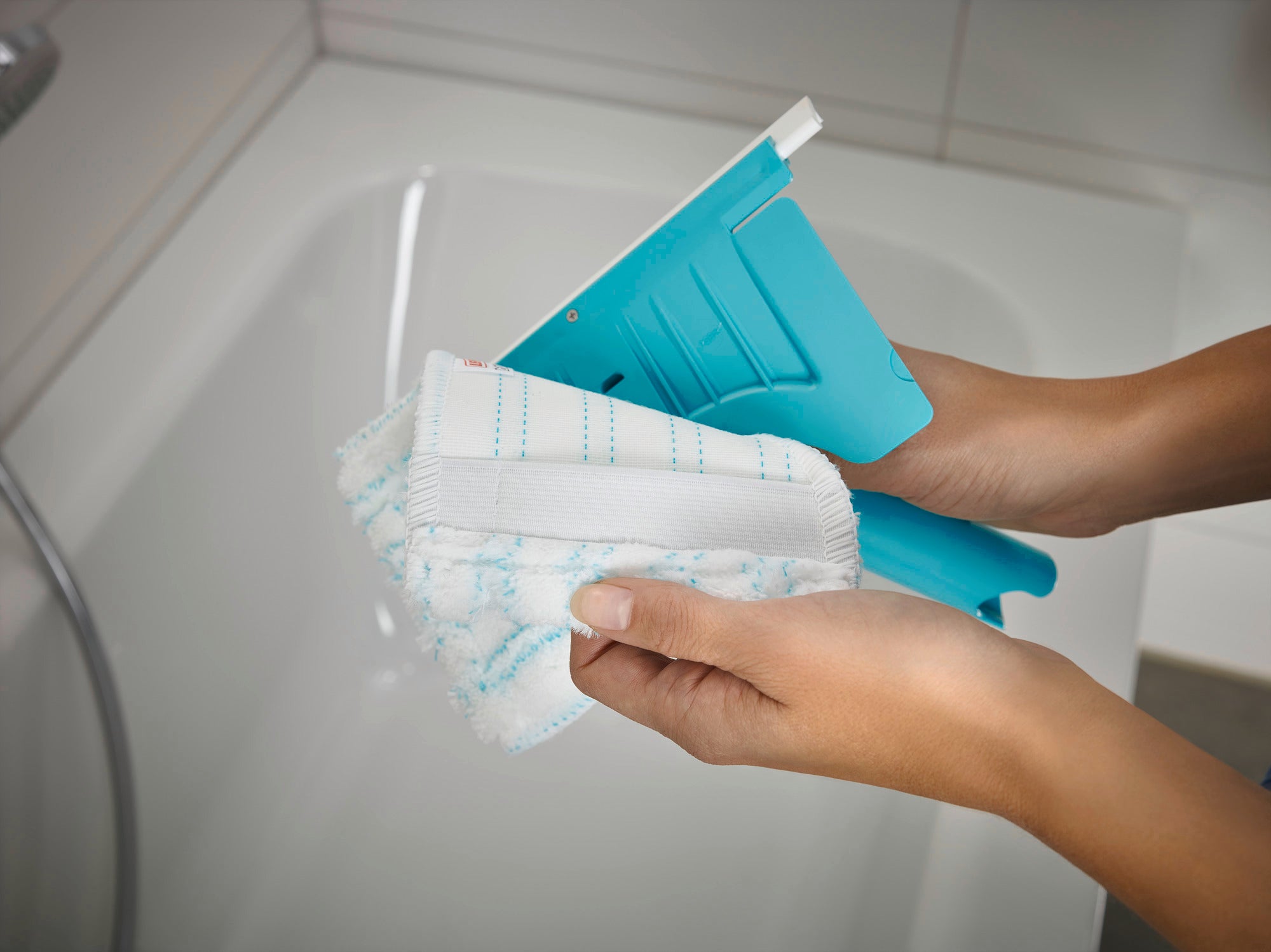 Flexipad tile and tub squeegee with telescopic handle in use. Two hands hold the squeegee over a bathtub. The squeegee has a blue pad and a white handle.