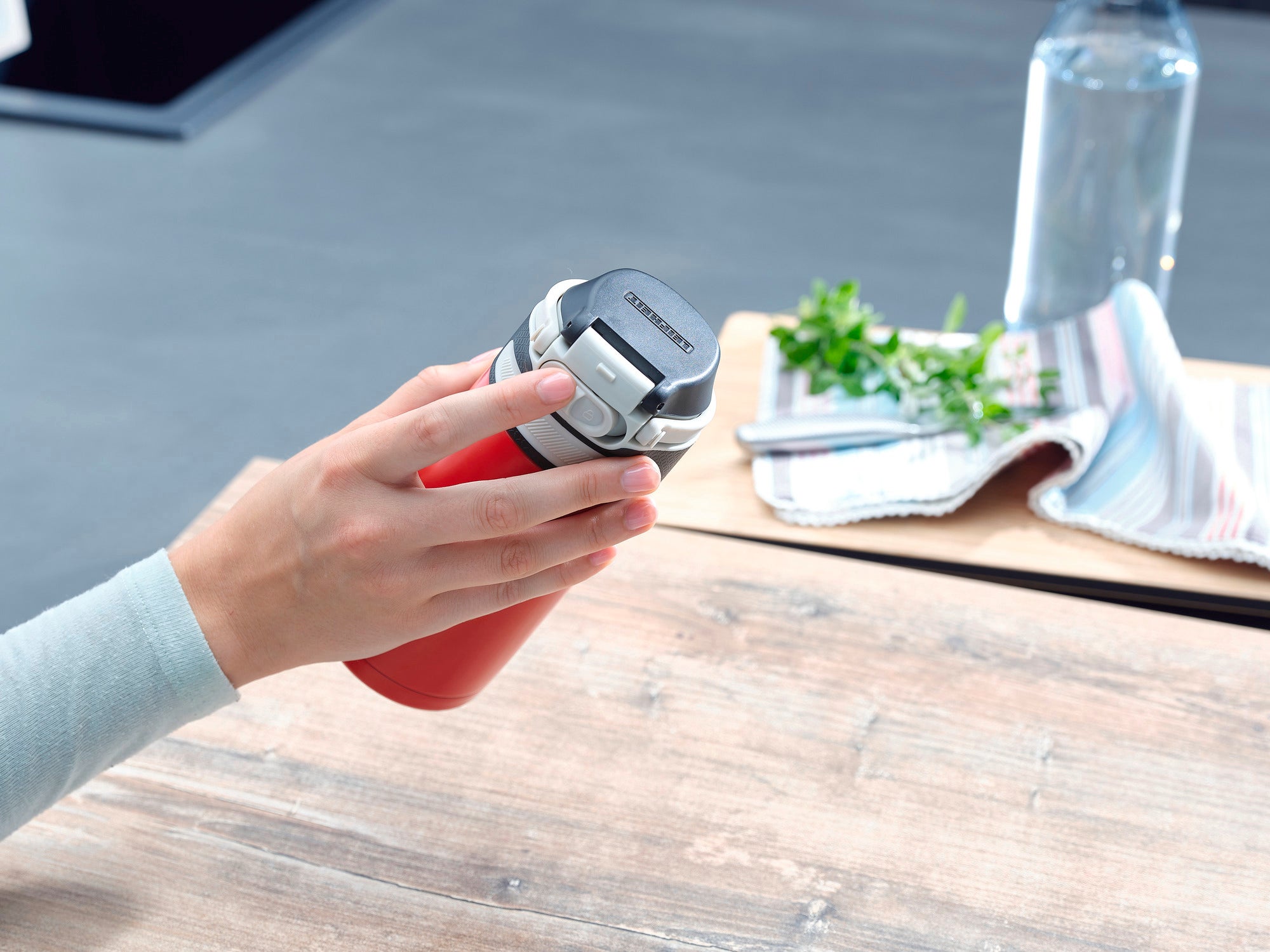 A red insulated mug with a flip lid is held in one hand. A tea towel, fresh herbs and a water bottle can be seen in the background.