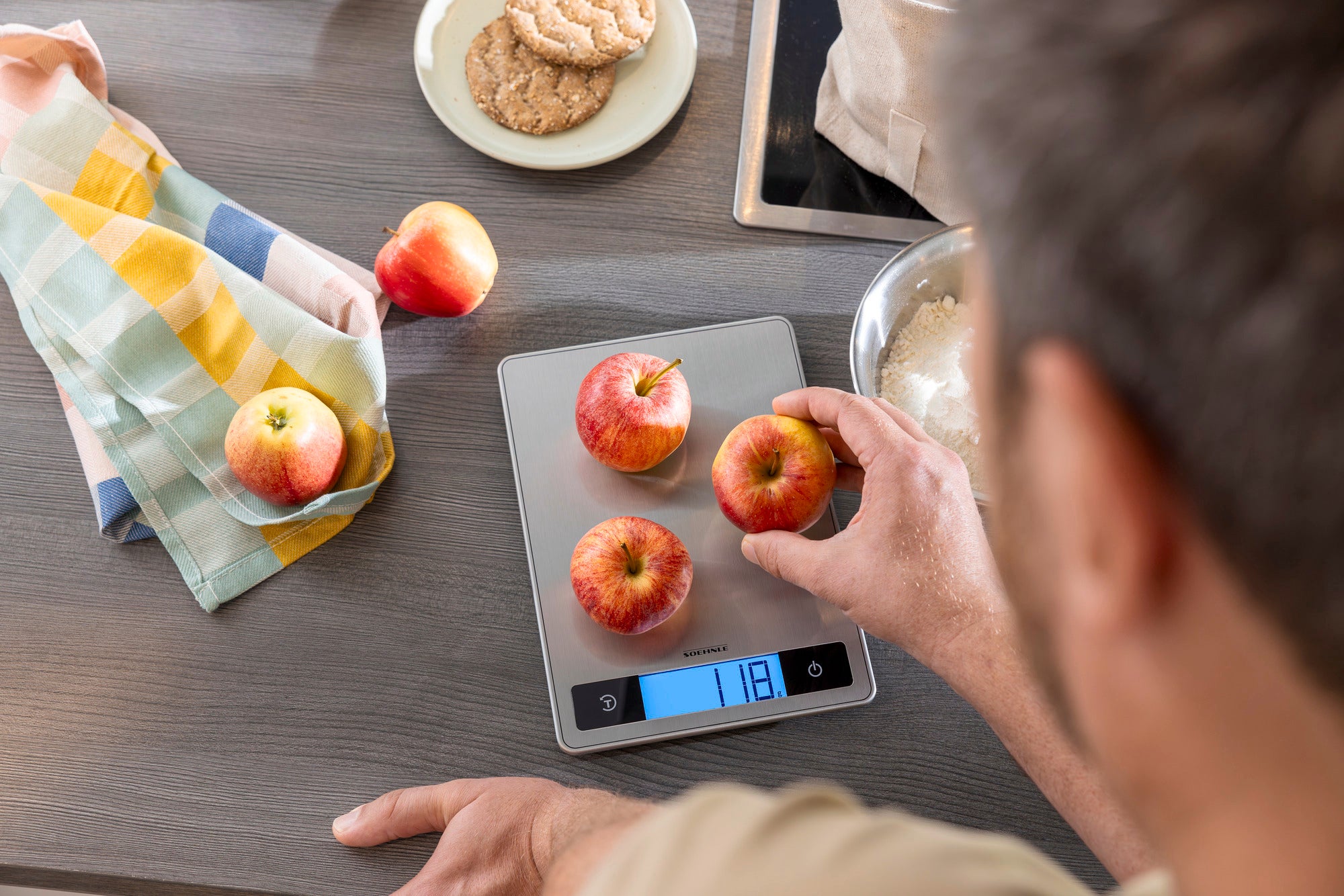 Een digitale keukenweegschaal die appels weegt. De weegschaal geeft een gewicht van 118 gram aan. Op de achtergrond zie je een theedoek, koekjes en een kom bloem.