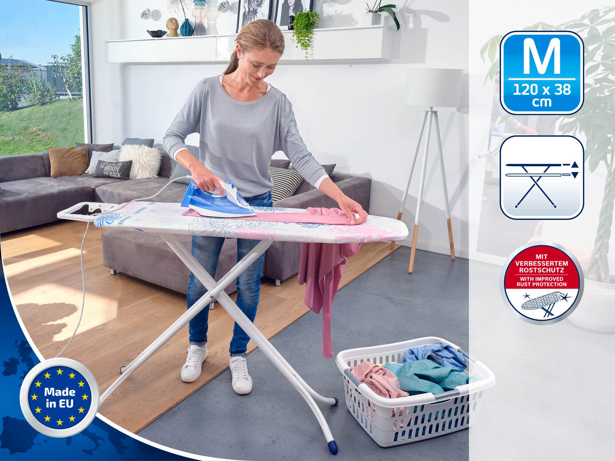 Une femme repasse une chemise rose sur une table à repasser dans un salon moderne. La table mesure 120 x 38 cm et est de construction robuste. Un panier à linge se trouve à côté.