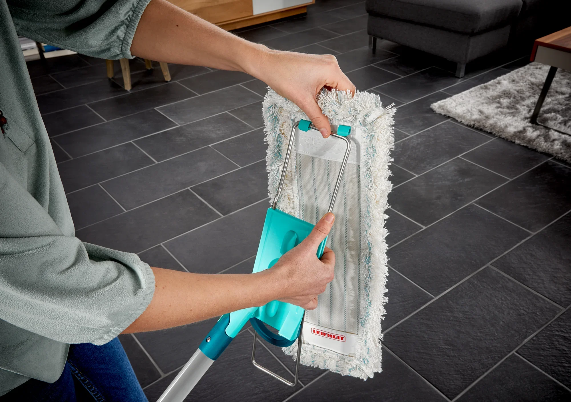 Eine Person benutzt den Bodenwischer Profi XL, um einen dunklen Fliesenboden zu reinigen. Der Wischer hat einen Baumwollbezug und einen stabilen Griff für effektive Reinigung.
