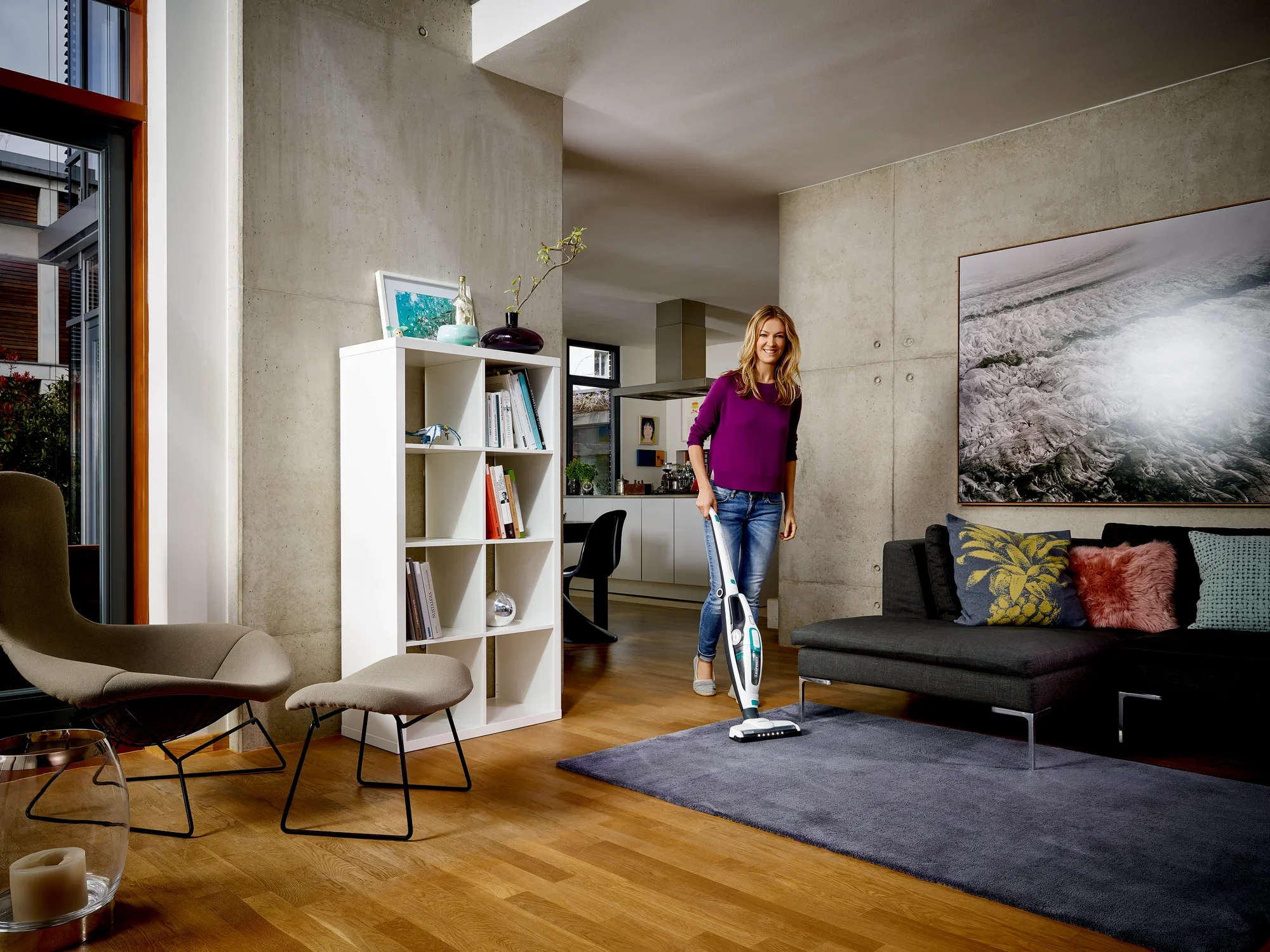 Une femme utilise l'aspirateur Regulus PowerVac 2 en 1 dans un salon moderne. La pièce est élégamment meublée avec un canapé gris, une bibliothèque et des œuvres d'art au mur.