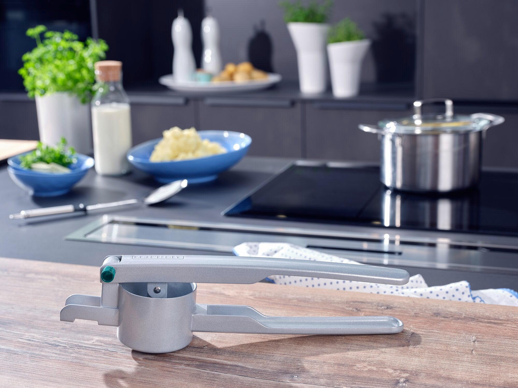 Eine Kartoffel-Spätzlepresse aus Metall liegt auf einer modernen Küchenarbeitsplatte. Im Hintergrund sind Zutaten wie Kartoffeln, Milch und Kräuter zu sehen.