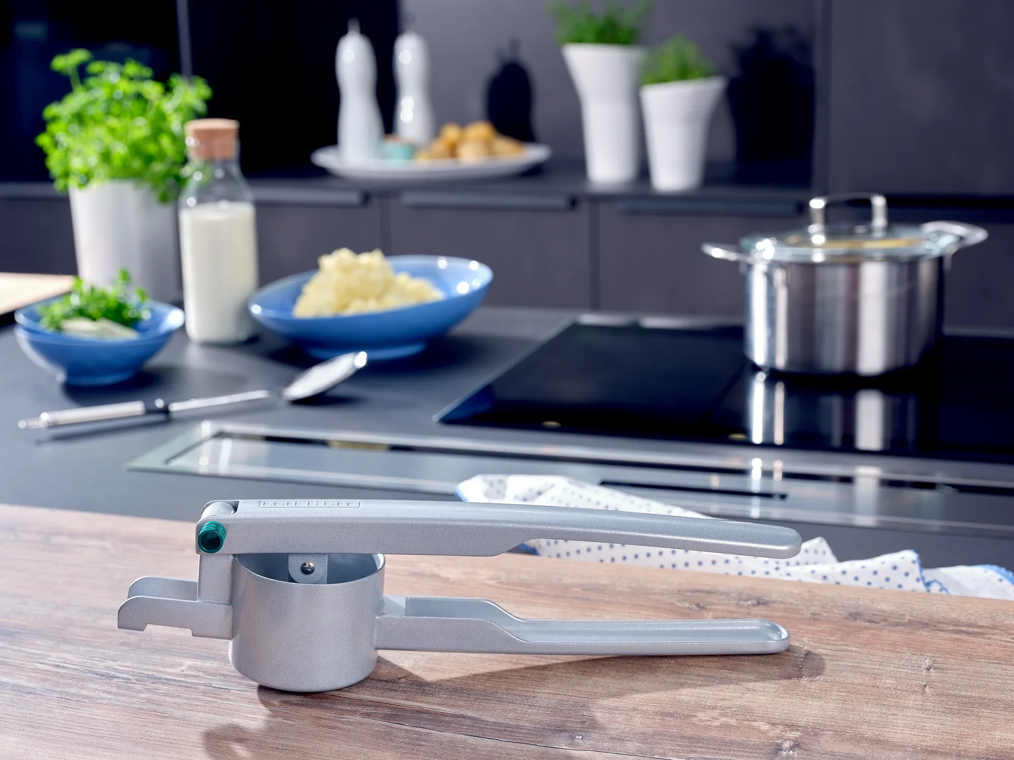 Metalowa prasa do ziemniaków leży na nowoczesnym blacie kuchennym. W tle widoczne są składniki takie jak ziemniaki, mleko i zioła.